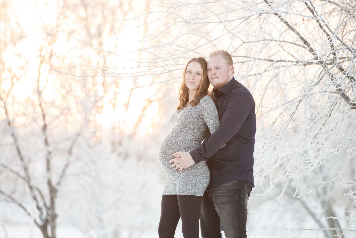 Pregnant couple embracing outdoors in snowy winter landscape with frost-covered trees and soft golden sunlight.