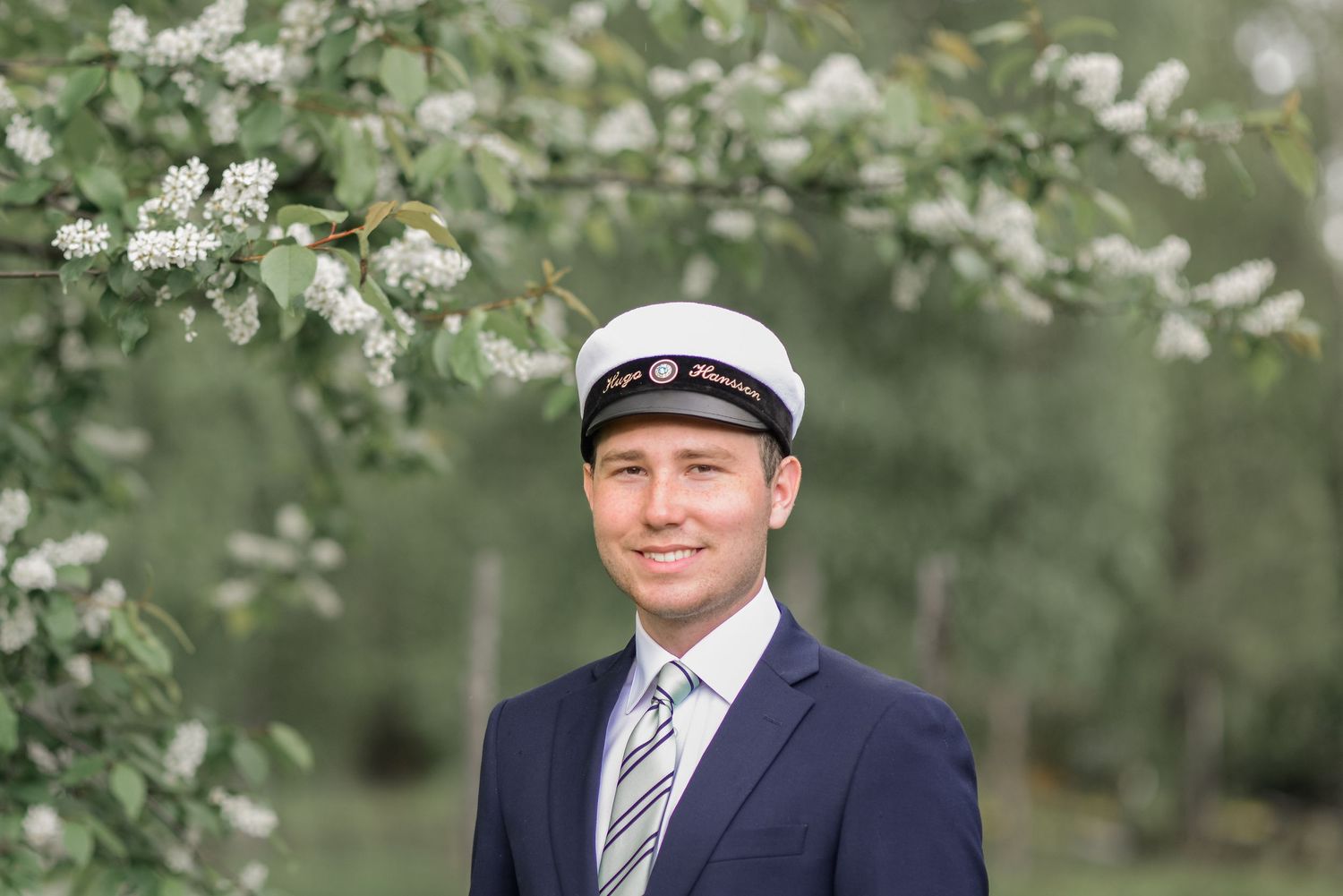 A person in formal attire and captain's hat smiles in front of blooming spring trees.