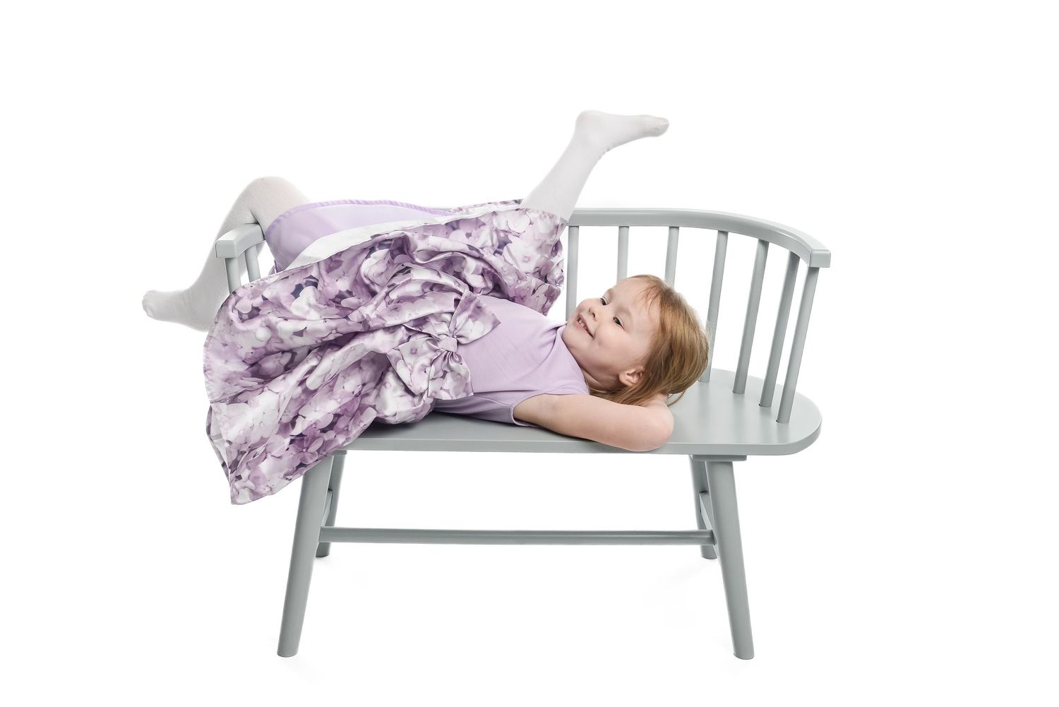 Child lying on gray bench in flowing dress against white background.