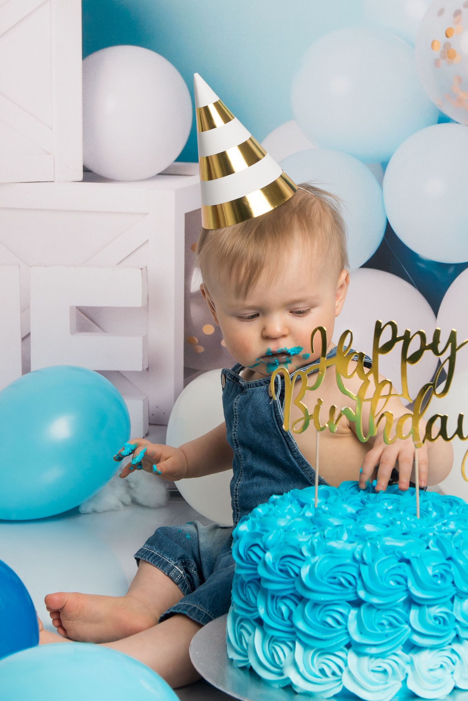 A bright blue birthday cake with rosette frosting and gold 'Birthday' cake topper in a festive balloon setting.