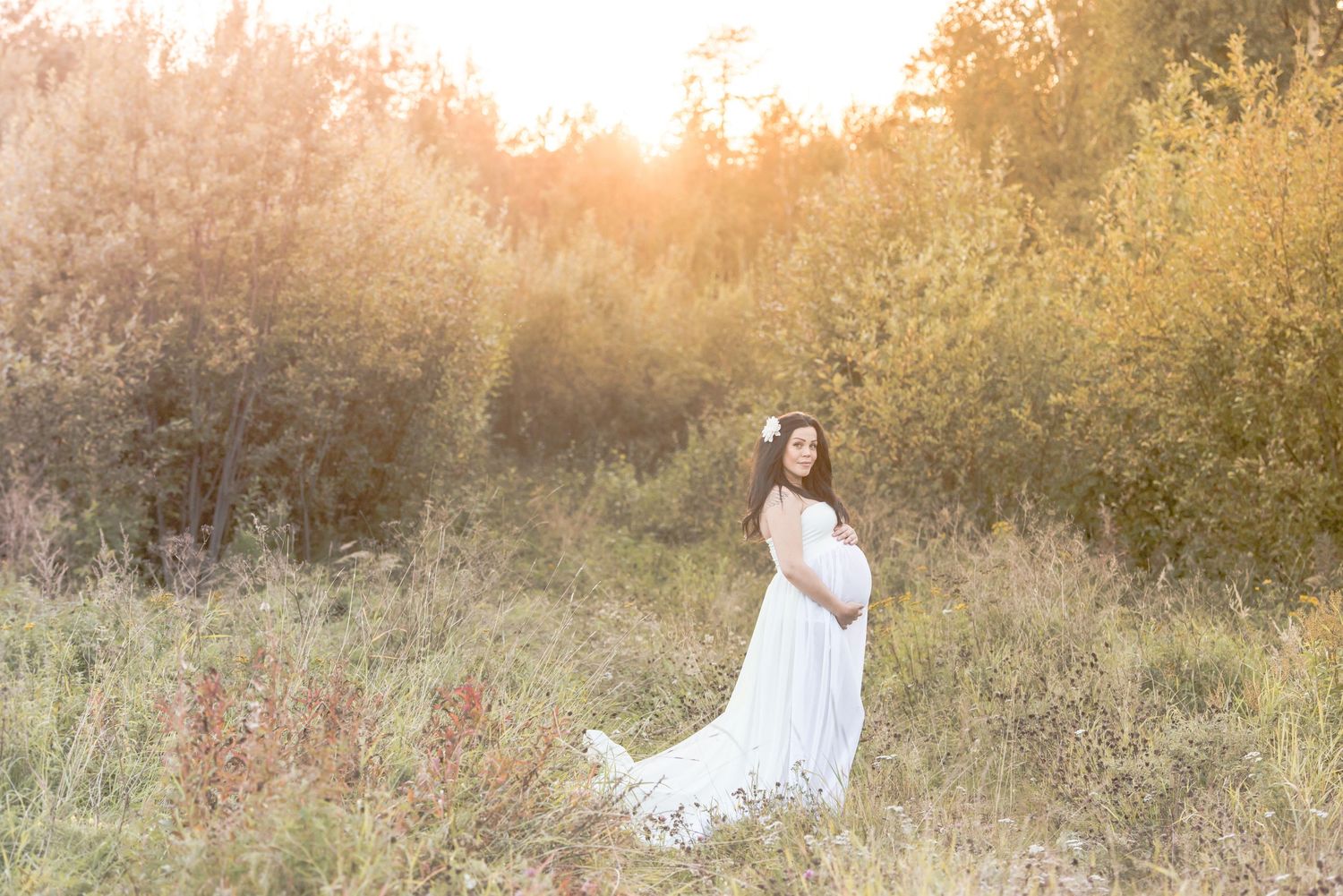 Pregnant woman in white gown and floral crown poses in golden sunlit meadow for maternity photography session.