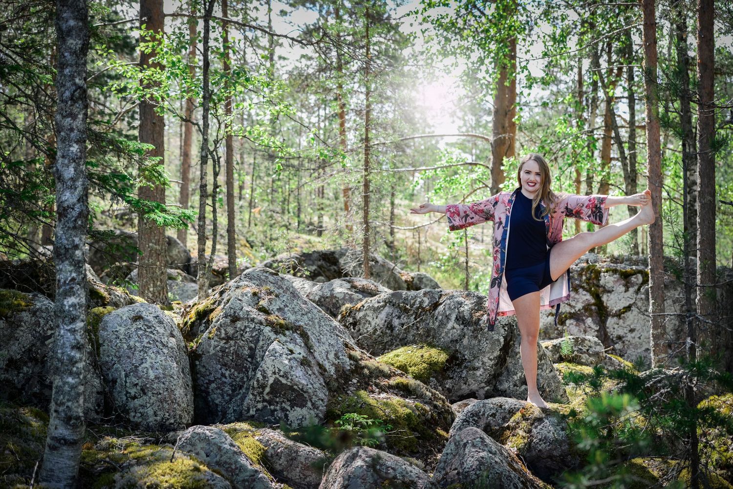 Young woman balancing on rocks in sunlit forest wearing plaid shirt and shorts.