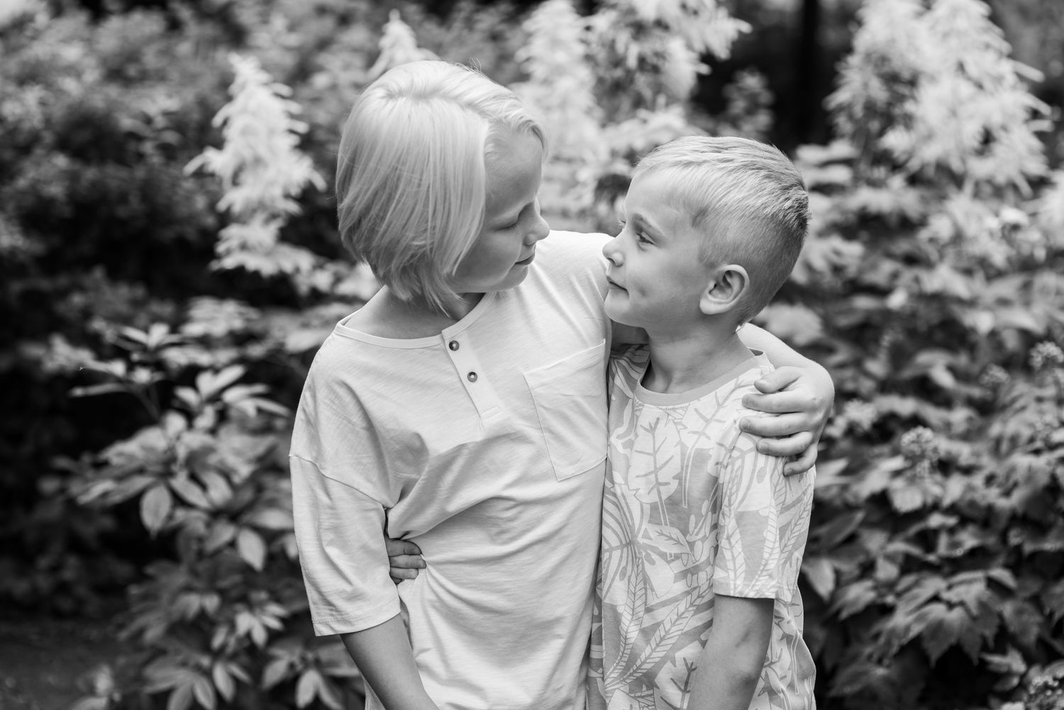Black and white photo of a mother and young son sharing a tender moment outdoors among lush garden foliage.