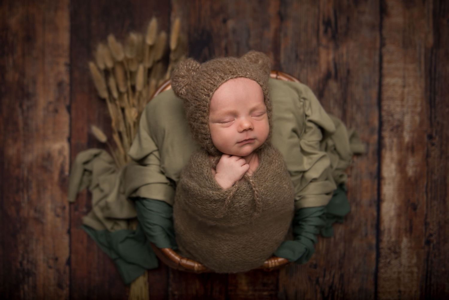 Sleeping newborn wrapped in green and brown fabrics on wooden backdrop.
