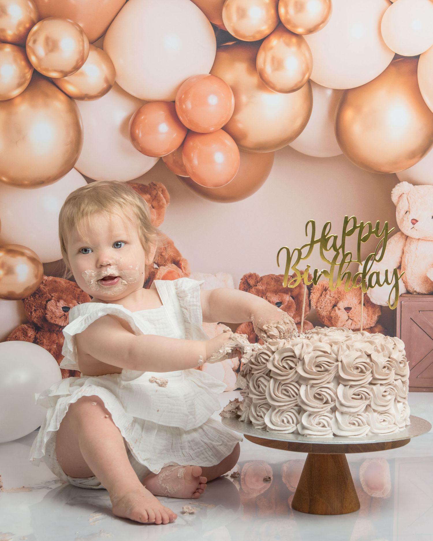 A decorated cake sits on a wooden stand with rose gold balloon backdrop for a celebration.