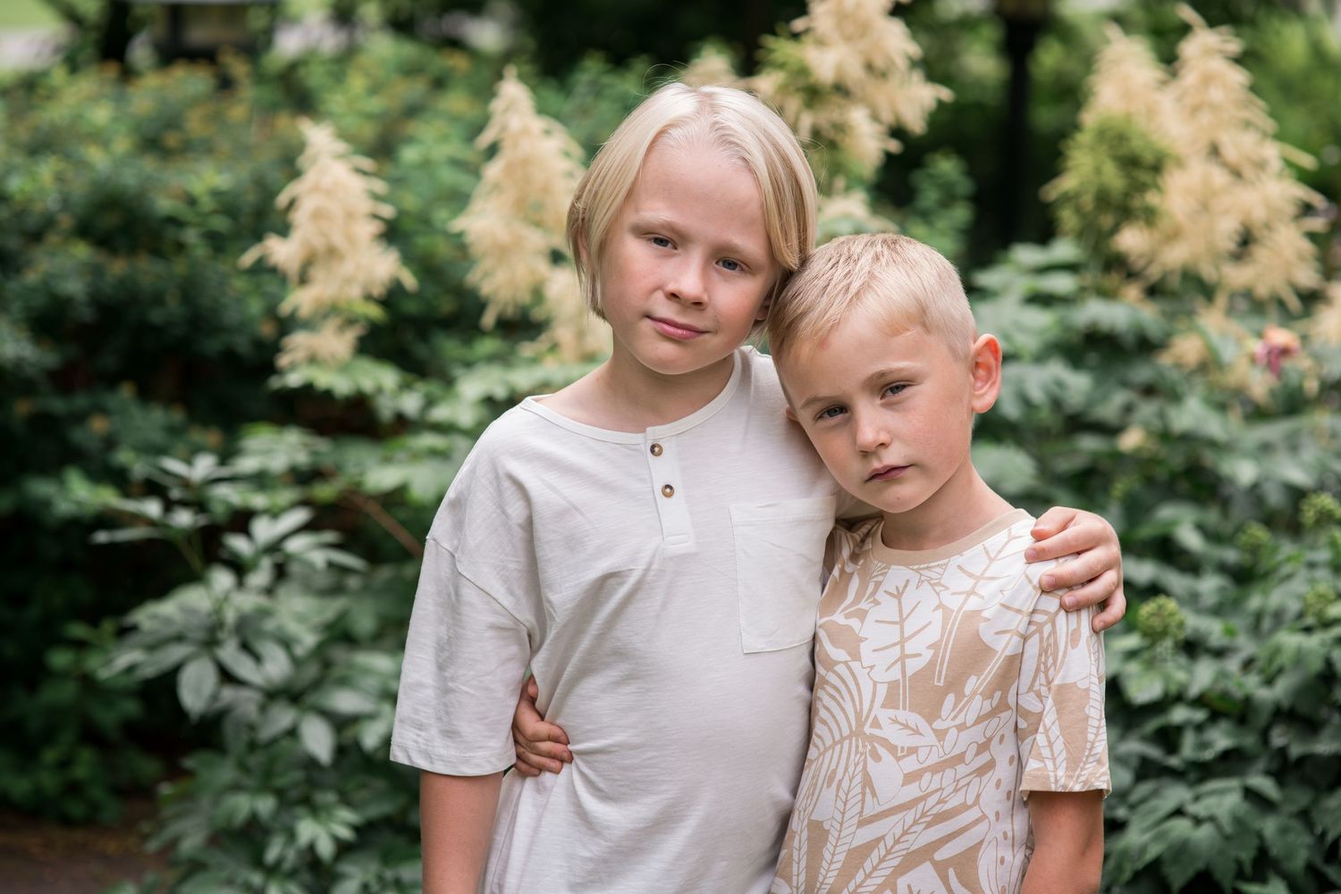 Two blonde boys in neutral tones pose together outdoors among lush green foliage and cream flowers.