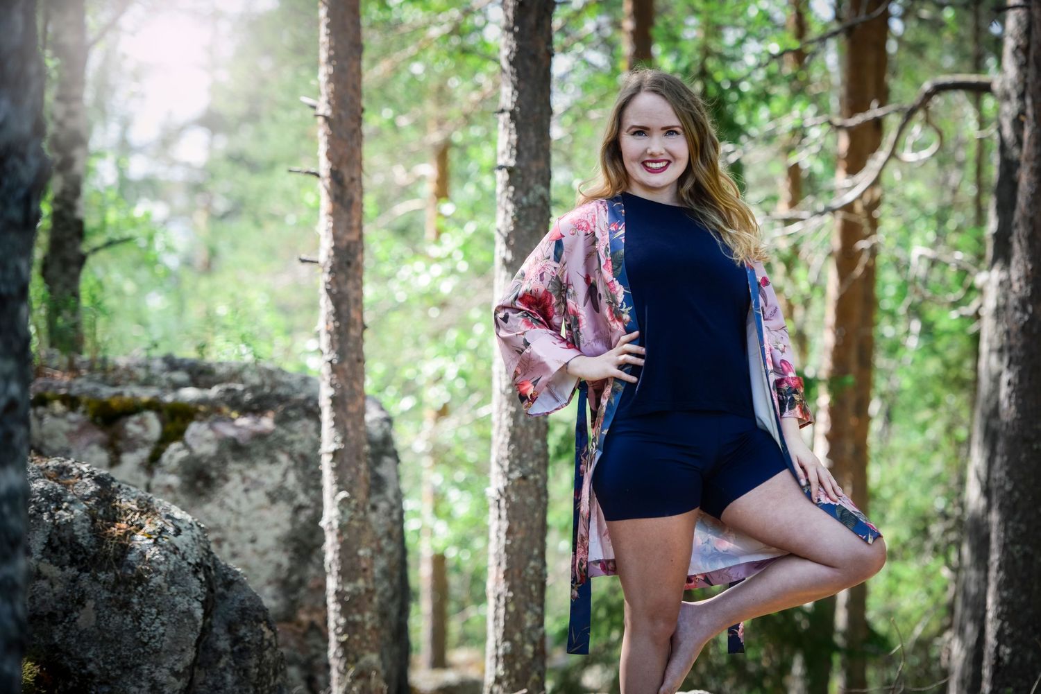 Smiling woman in navy romper and floral kimono posing gracefully on one leg in a forest setting.