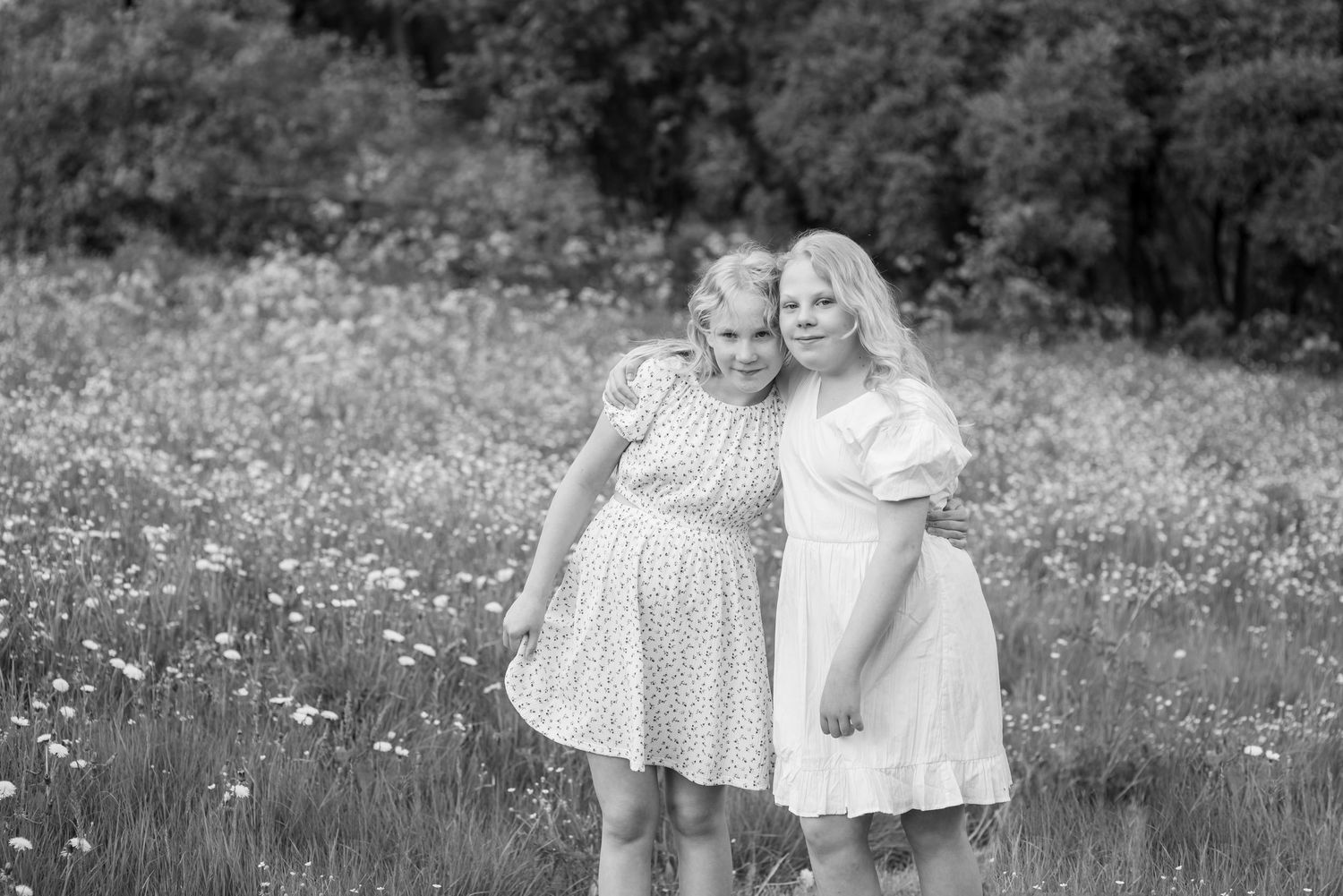Two women in floral dresses embrace in a wildflower meadow, captured in a timeless black and white portrait.
