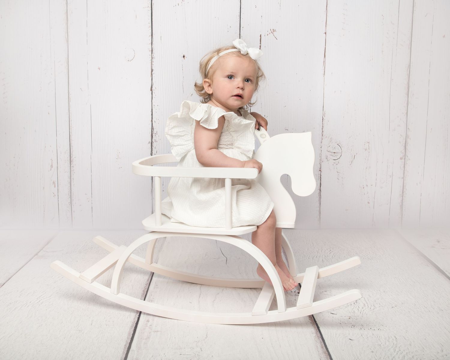 Adorable baby girl in white dress sitting on a white wooden rocking horse against a white background.