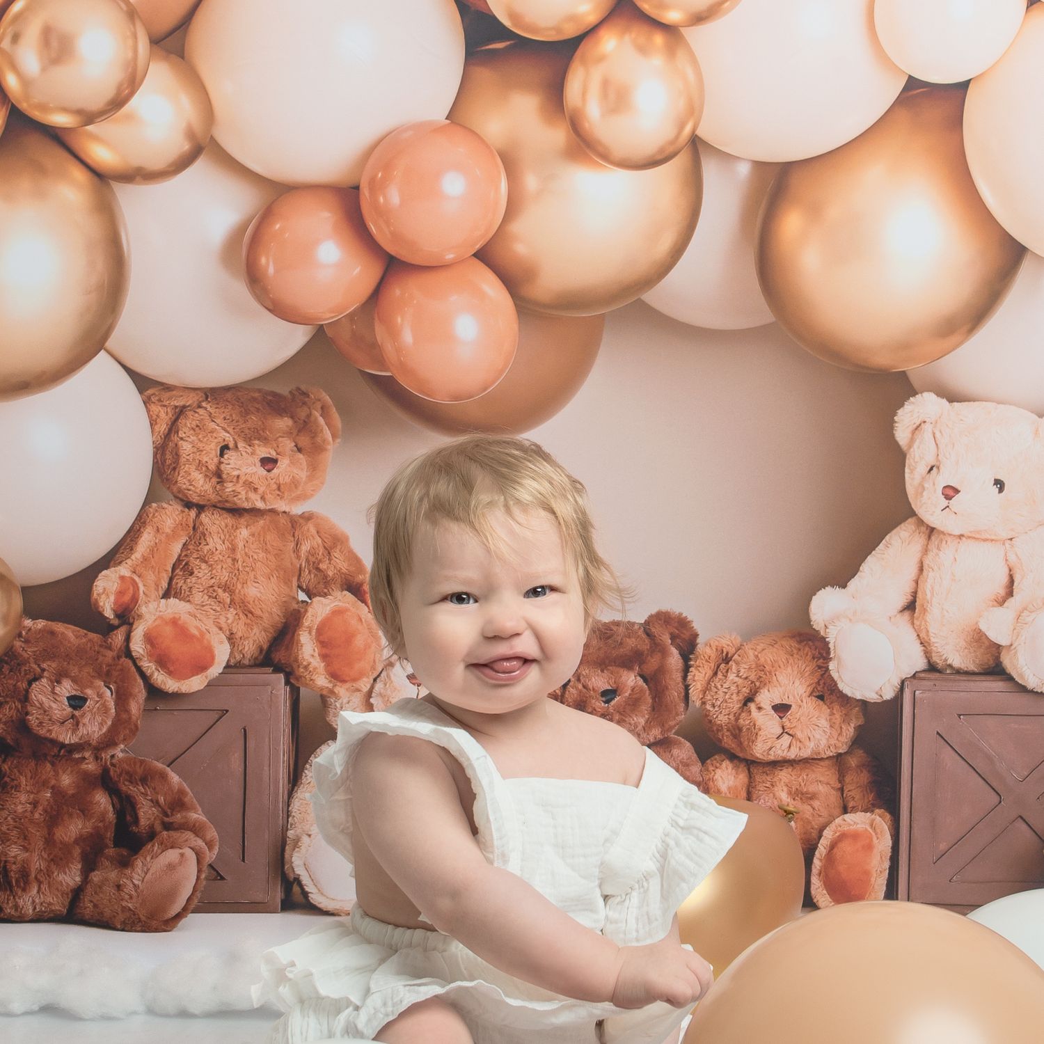 A baby in white outfit sits among rose gold balloons and teddy bear decorations.