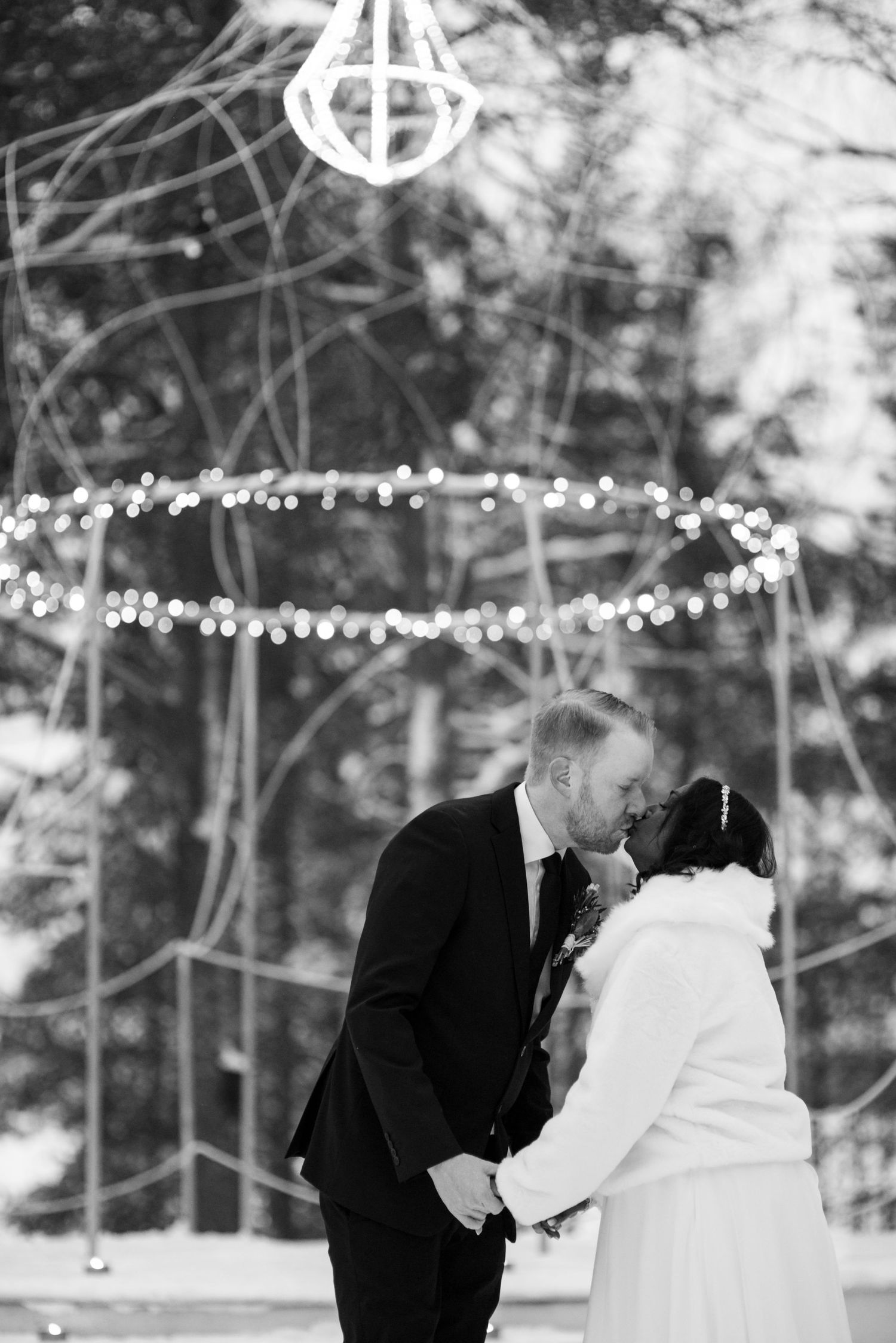 String lights twinkle above a romantic winter wedding moment in a forest setting.