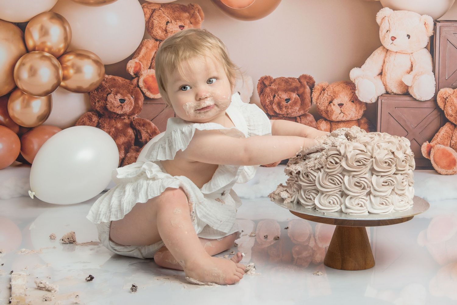 Sweet first birthday setup with pink balloons, teddy bears, and a baby in white dress against neutral backdrop.