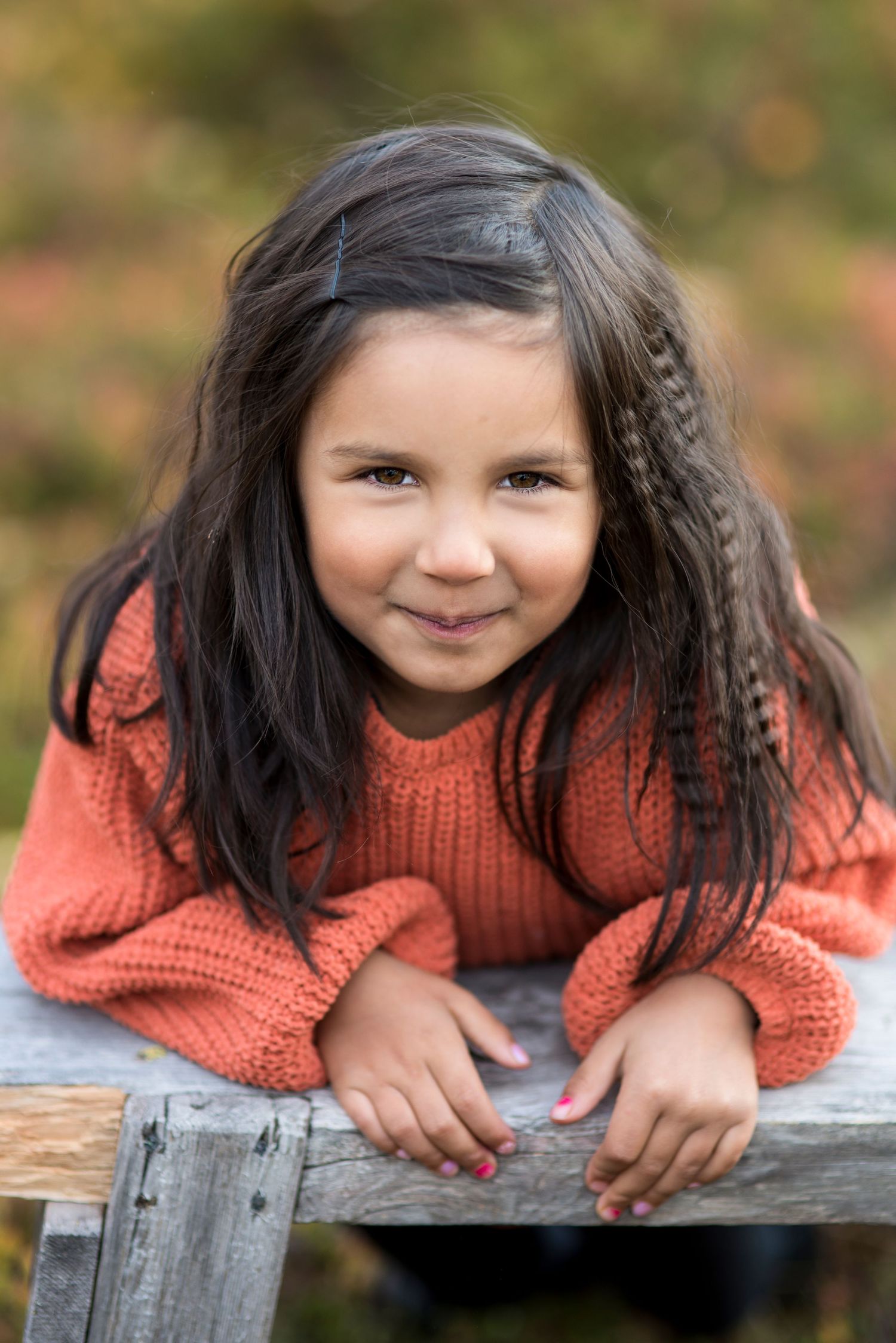 Young girl in orange knit sweater leaning on wooden fence, smiling outdoors with braided hair.