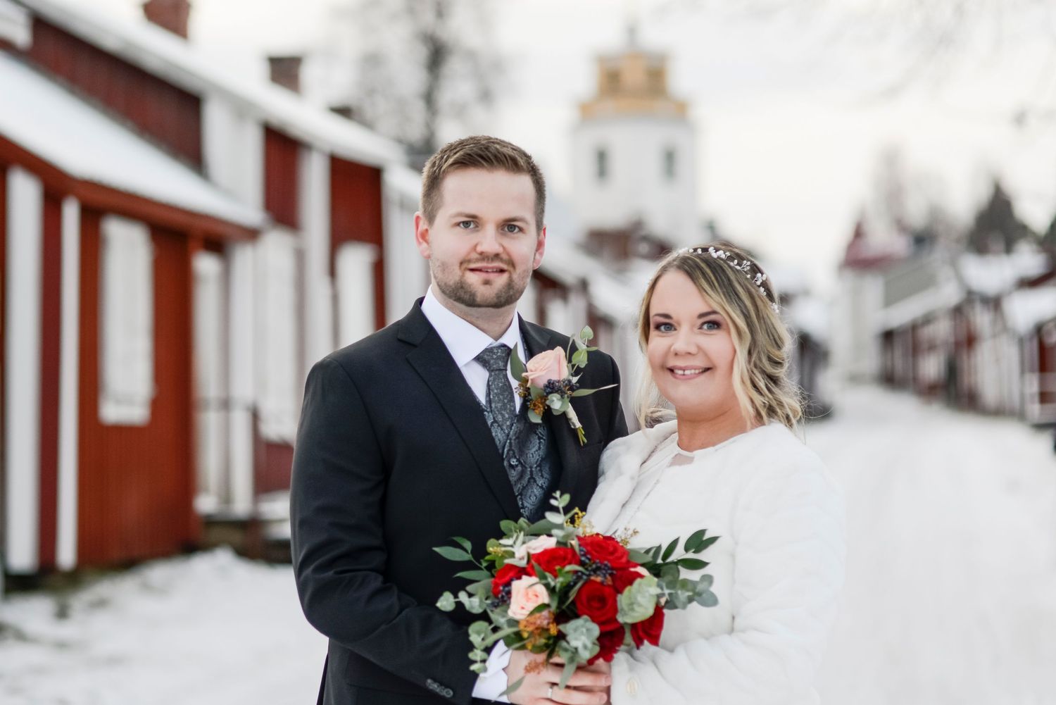 A winter wedding couple poses in the snow with red flowers against traditional red Swedish buildings.
