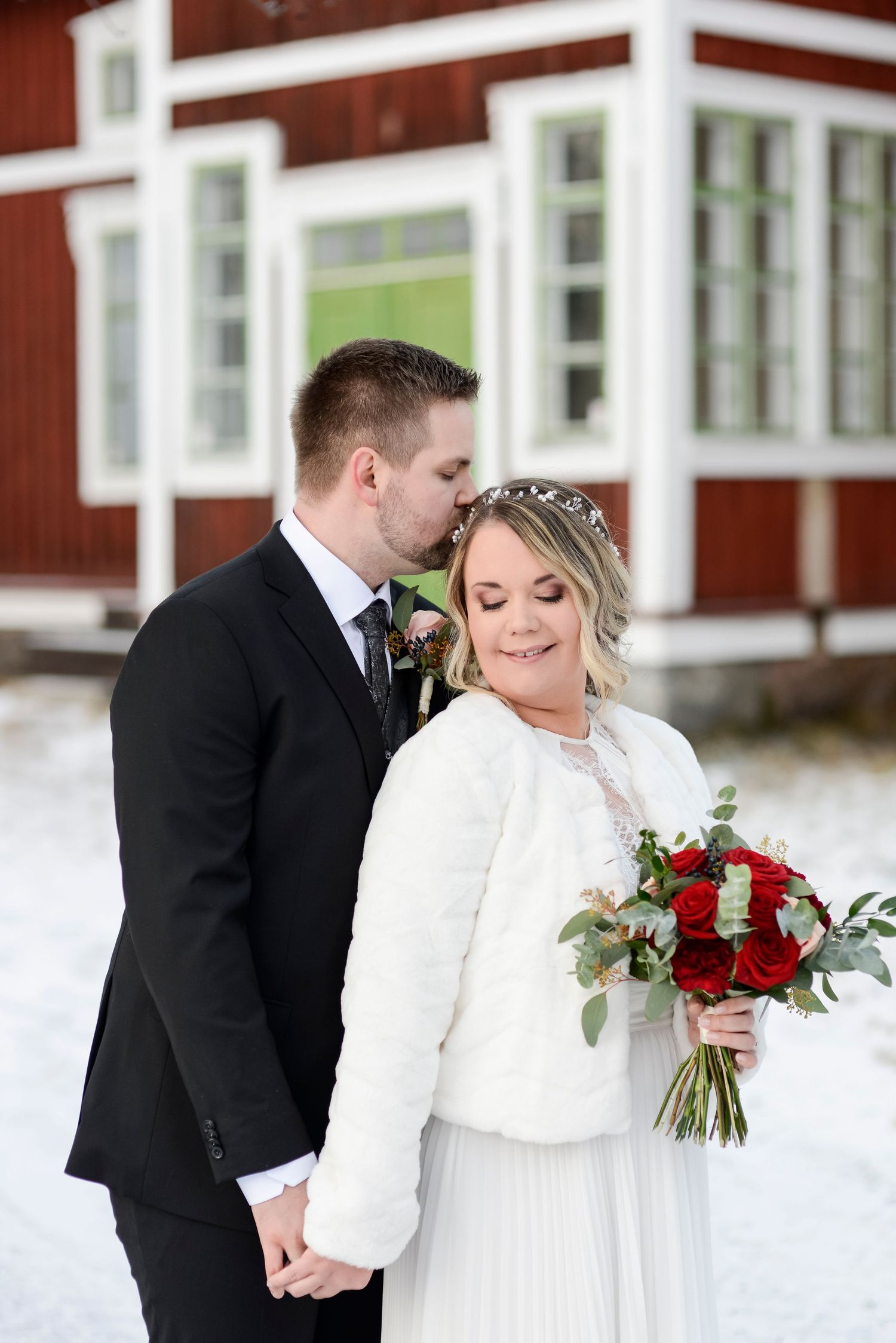 Winter wedding couple shares tender moment by red farmhouse with white trim.