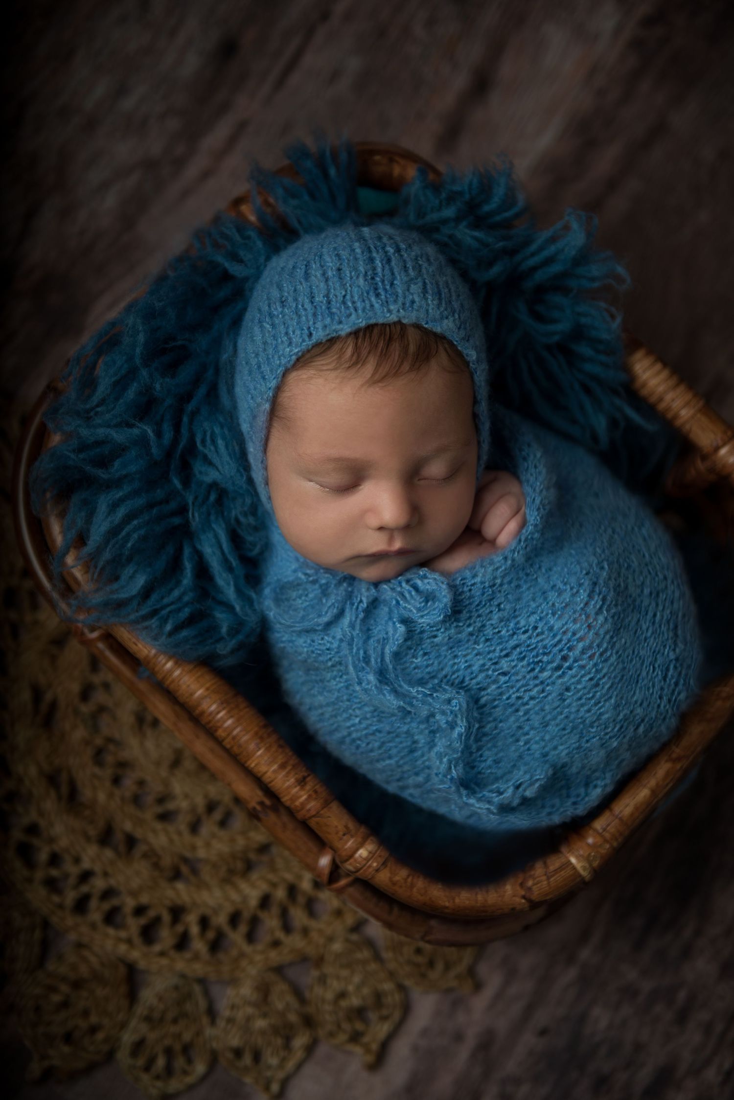 Newborn sleeping peacefully in blue knitted wrap and hat in wooden basket.
