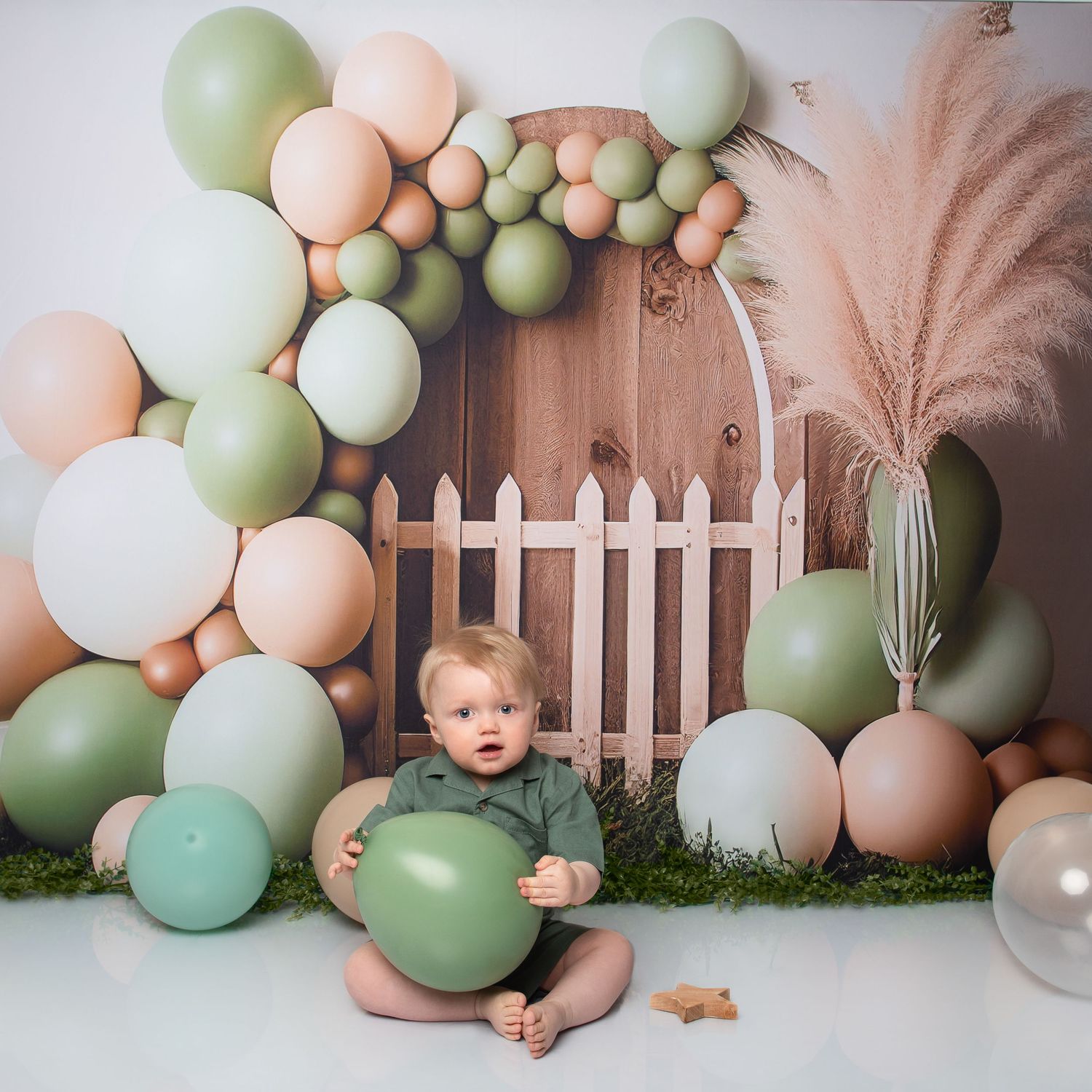 A baby sits on grass surrounded by sage green and peach balloons in a whimsical birthday photo setup with a wooden door backdrop.