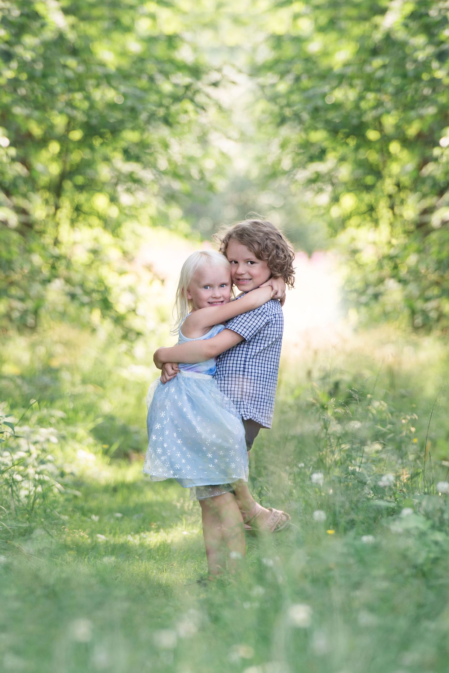 Two young children hugging in a lush green garden path, girl in blue polka dot dress embracing curly-haired boy.