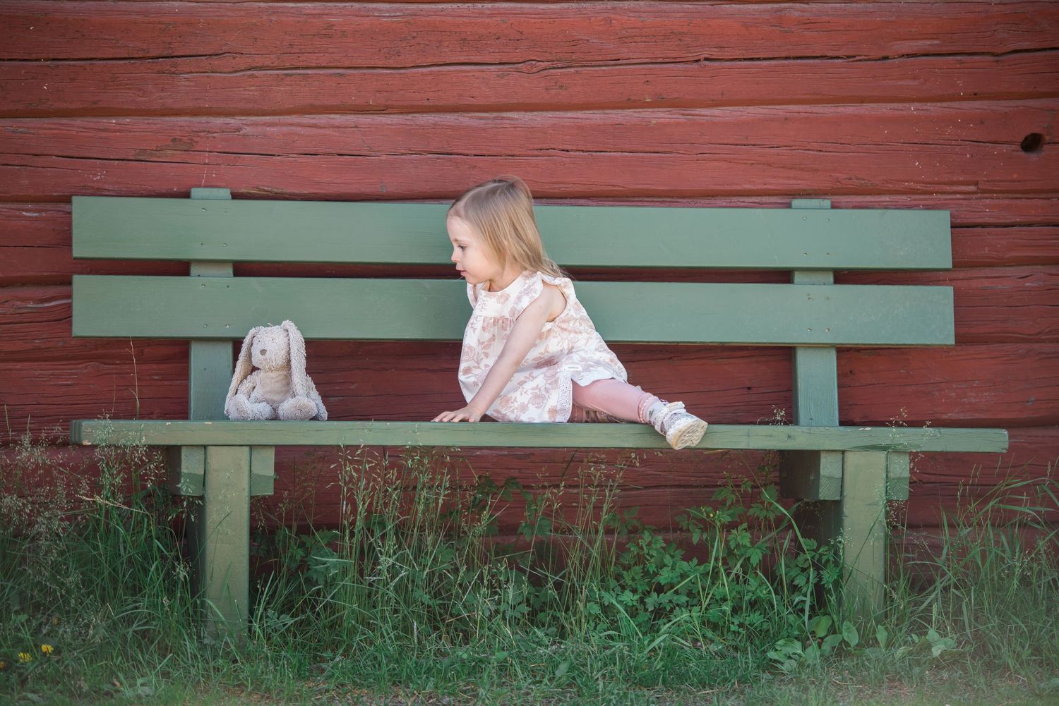 Young toddler girl sitting on a green wooden bench next to a stuffed bunny toy against a red barn wall.