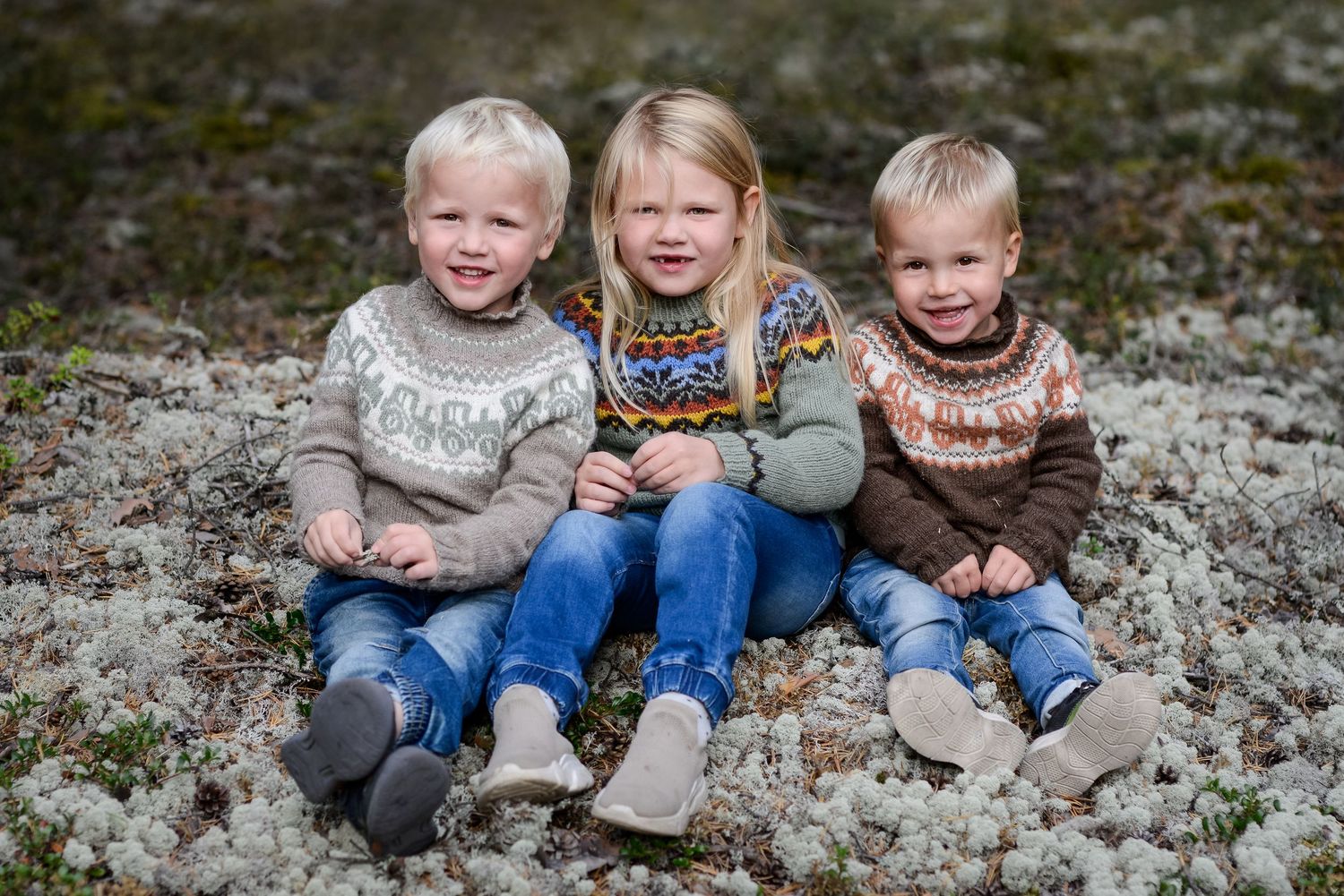 Three smiling young children in Nordic sweaters sitting together on mossy ground for a family portrait photo.