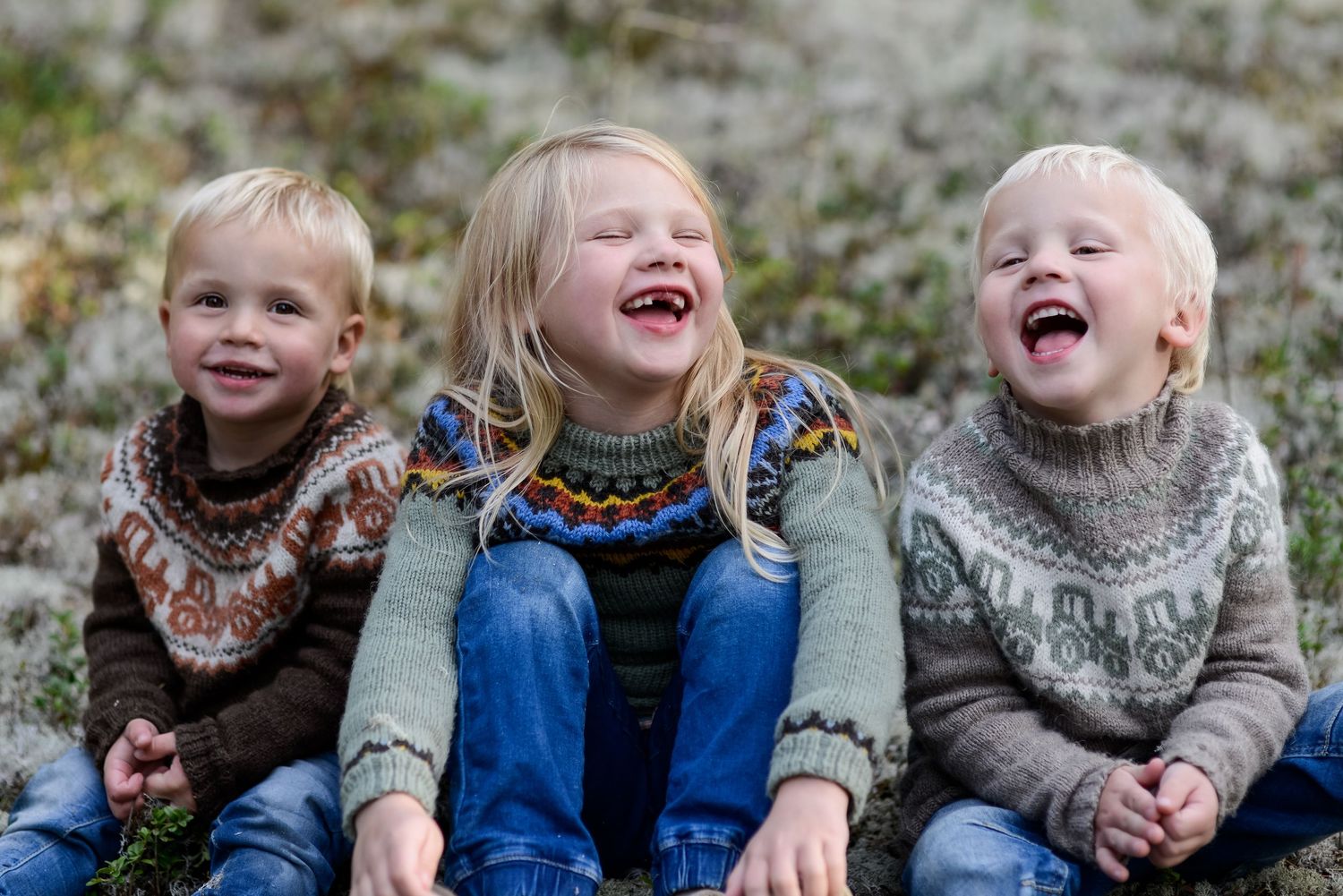 Three laughing children wearing Nordic sweaters sit together outdoors, sharing a joyful moment.