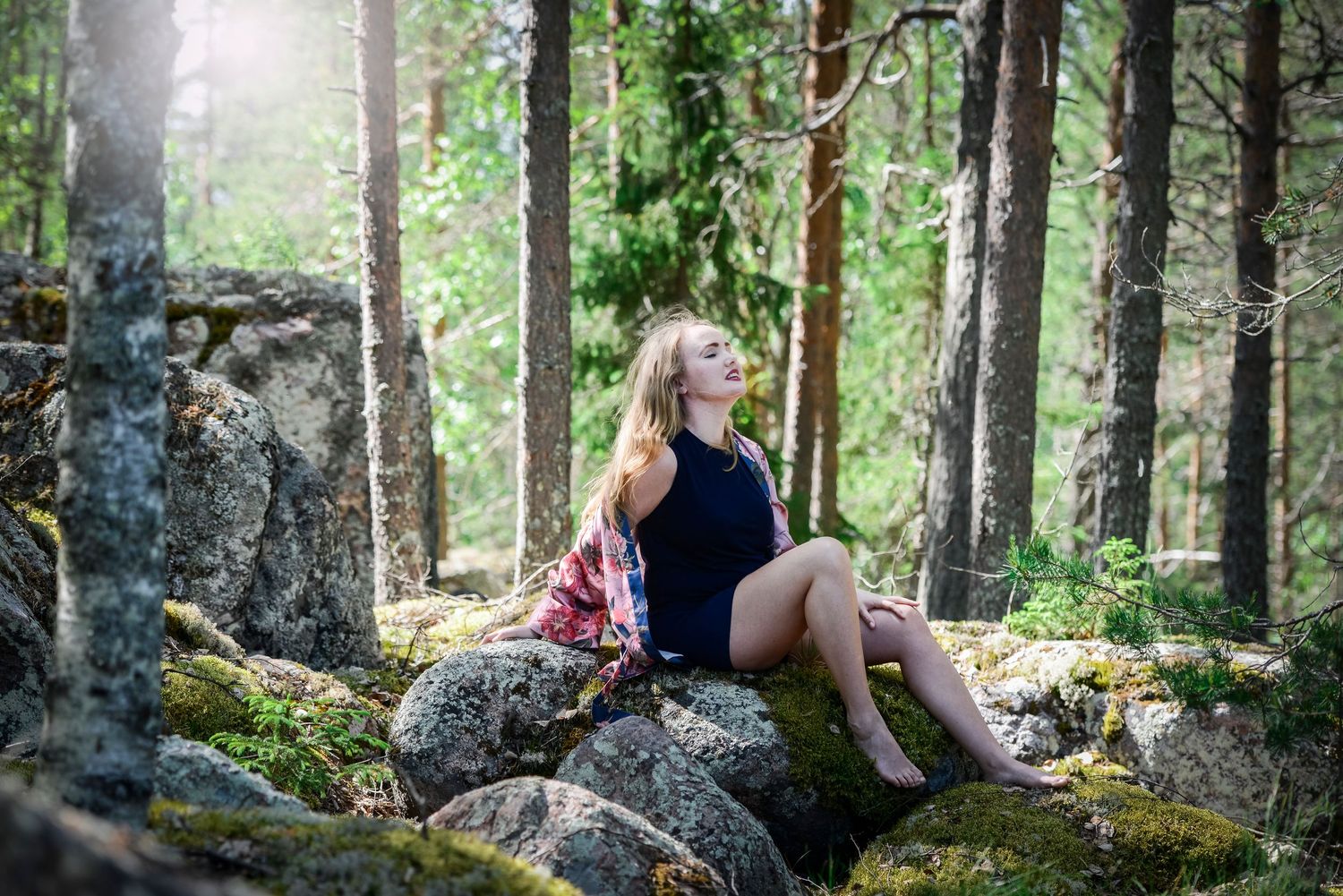 Woman sitting on mossy rocks in a sunlit forest, eyes closed, wearing a navy bodysuit and floral jacket.
