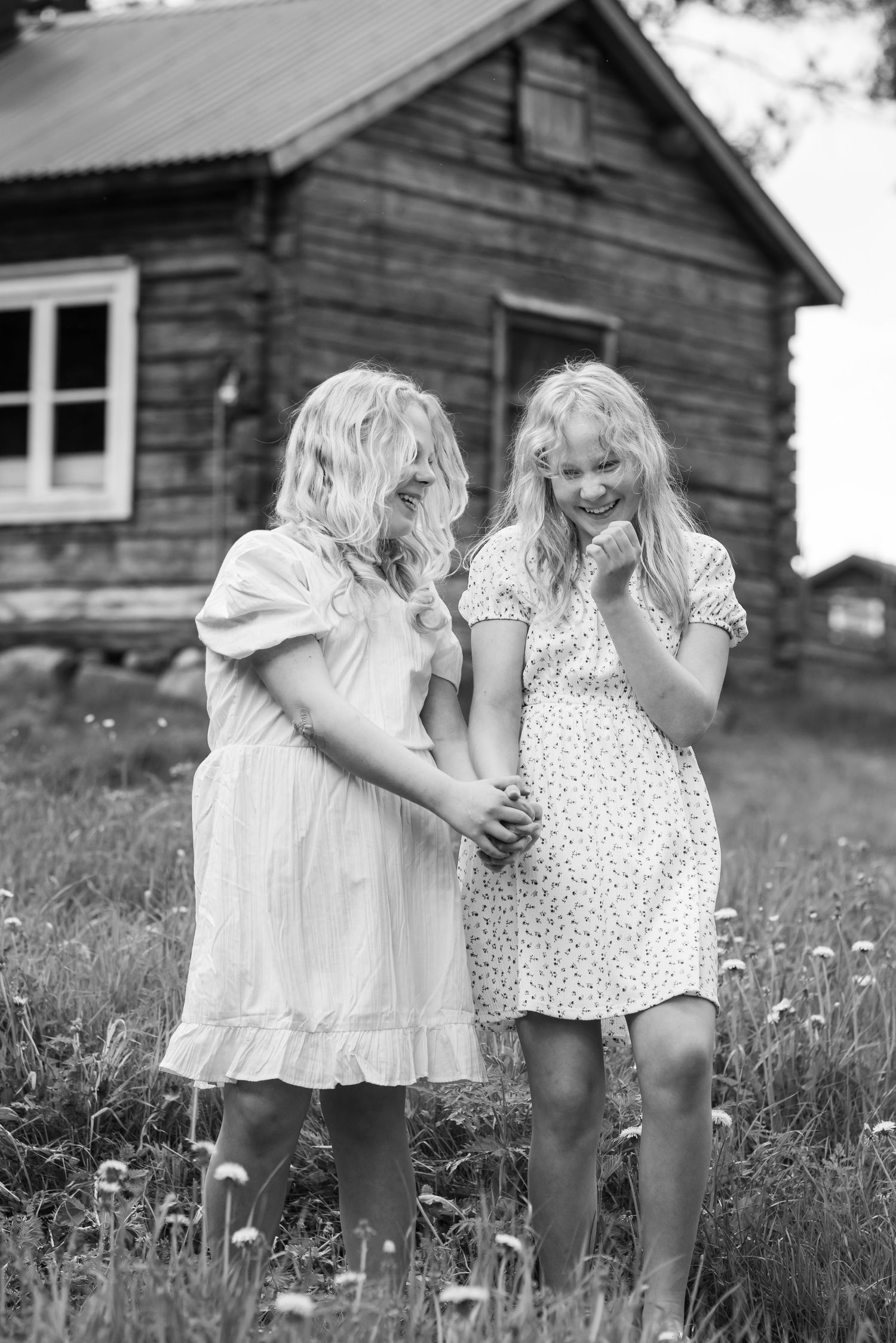 Two blonde girls in white dresses laugh together in a field beside a rustic wooden cabin, black and white photo.