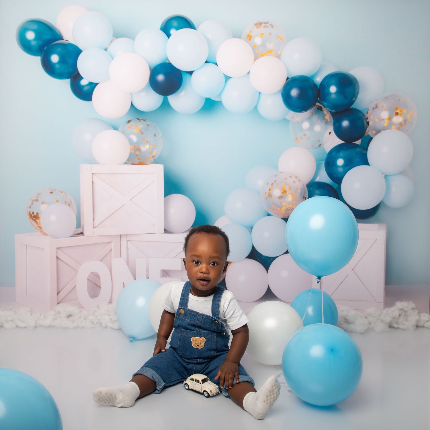 A baby boy in denim overalls sits on the floor surrounded by blue and white balloons at a first birthday celebration.