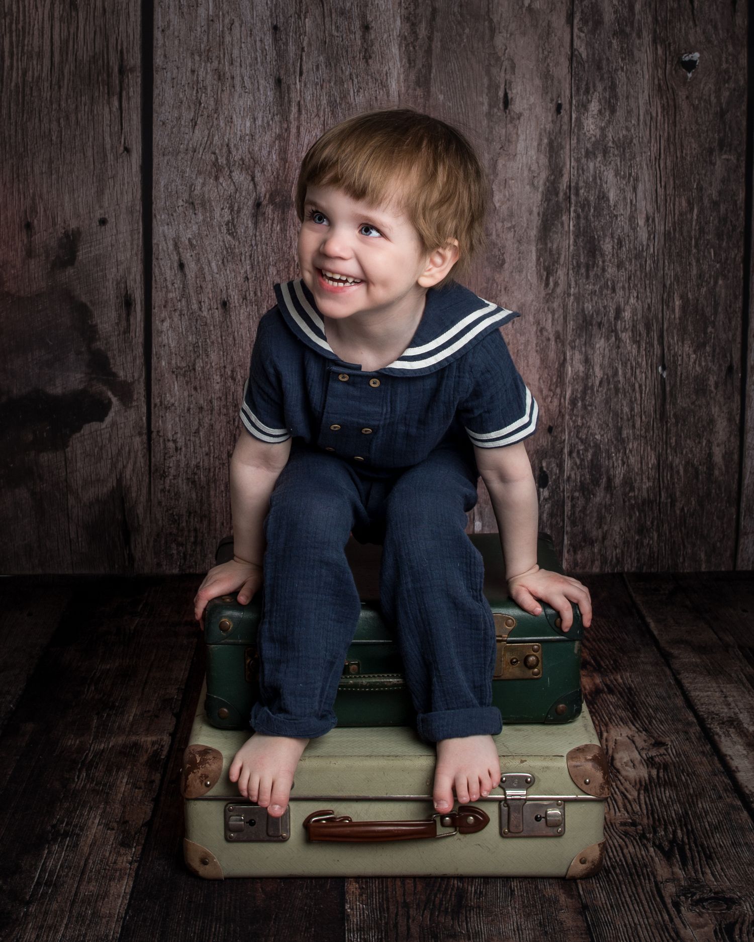 A smiling toddler boy in a navy sailor outfit sits barefoot on vintage stacked suitcases against a rustic wood backdrop.