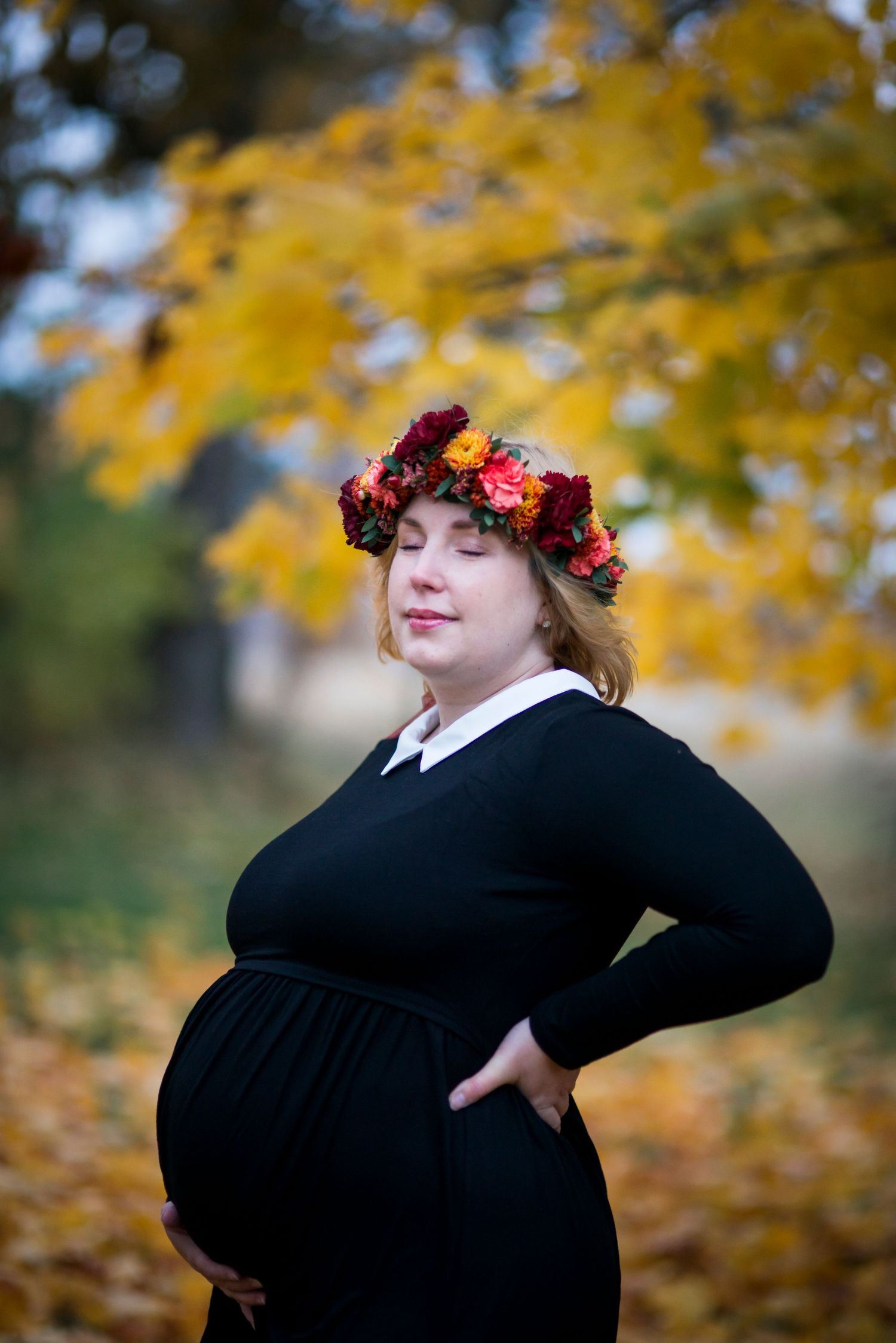 Pregnant woman in black dress and floral crown poses outdoors among golden autumn leaves.