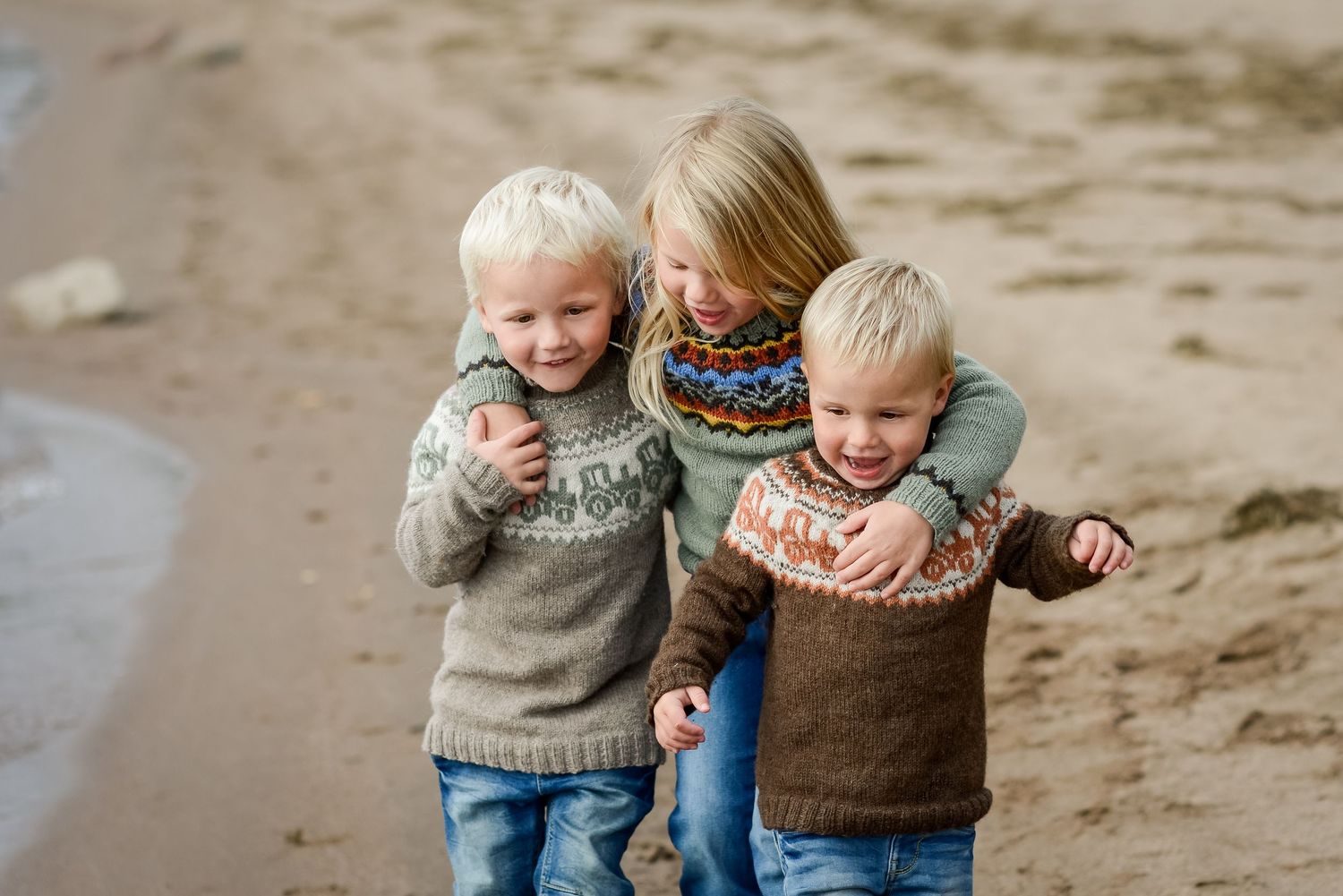 Three children in Nordic sweaters laugh and hug on a sandy beach, capturing a joyful sibling moment.