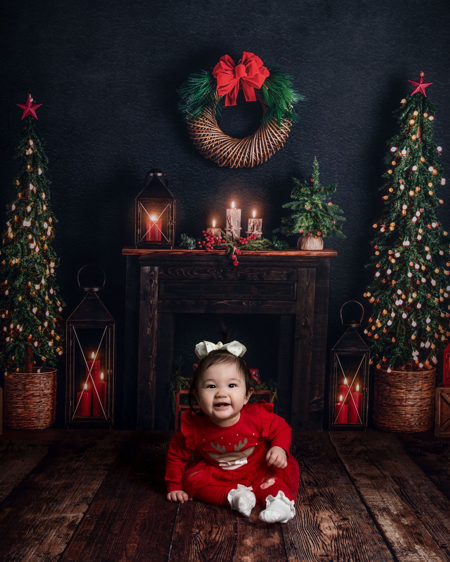 Cozy Christmas photo setup with decorated trees, red lanterns, and rustic wooden floor against dark backdrop.