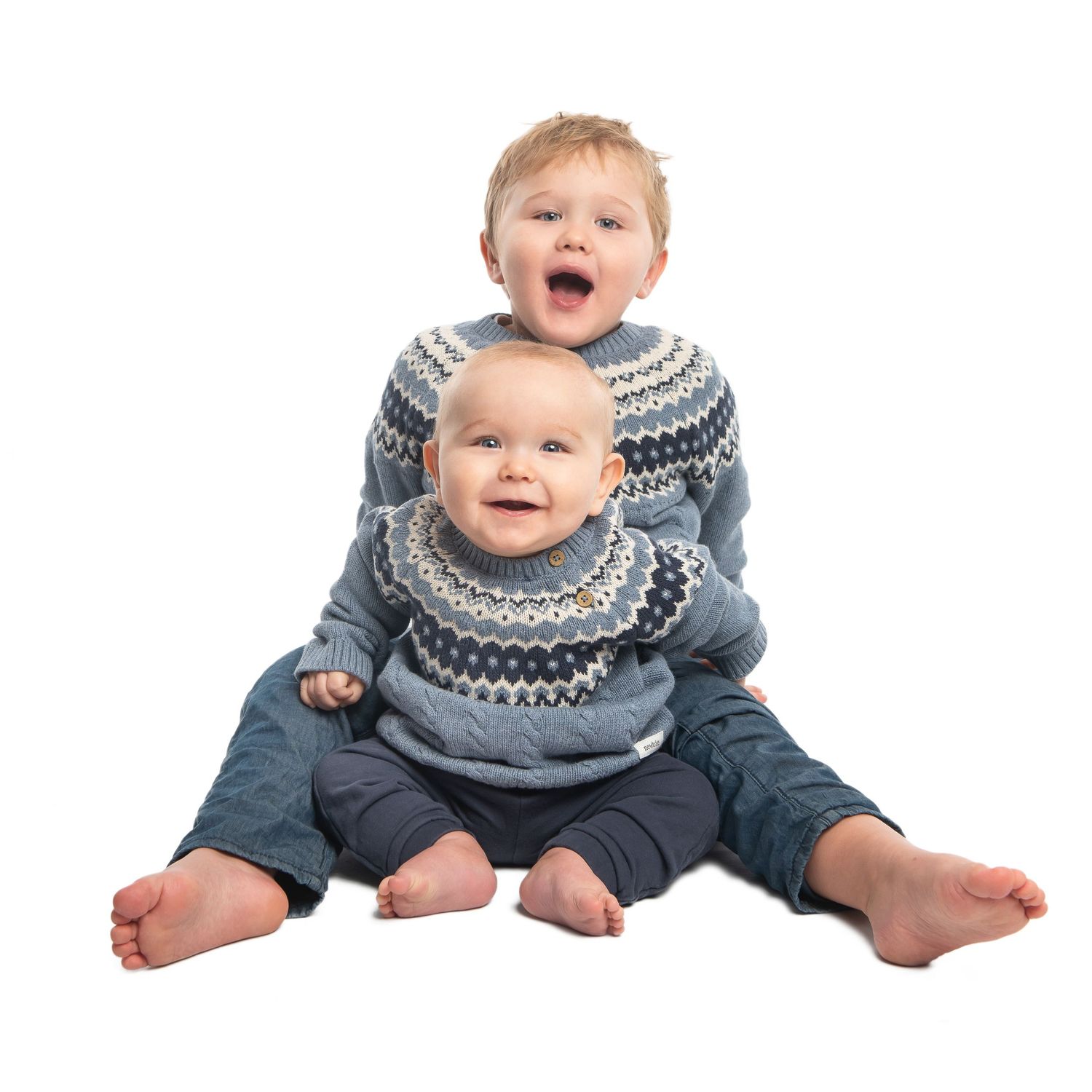 Baby wearing gray winter sweater sitting with legs spread on white surface.