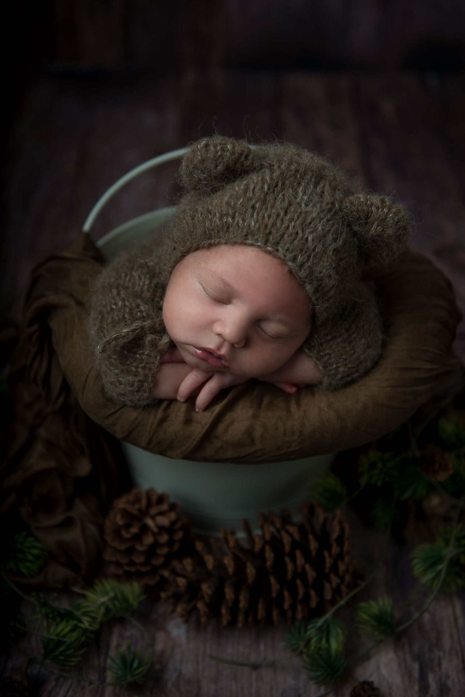 Sleeping baby in knit bonnet resting in vintage metal bowl with rustic pine cone decorations.