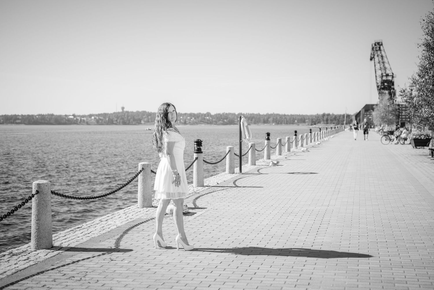 Woman in white dress standing on sunny waterfront promenade with rope bollards and crane in background.