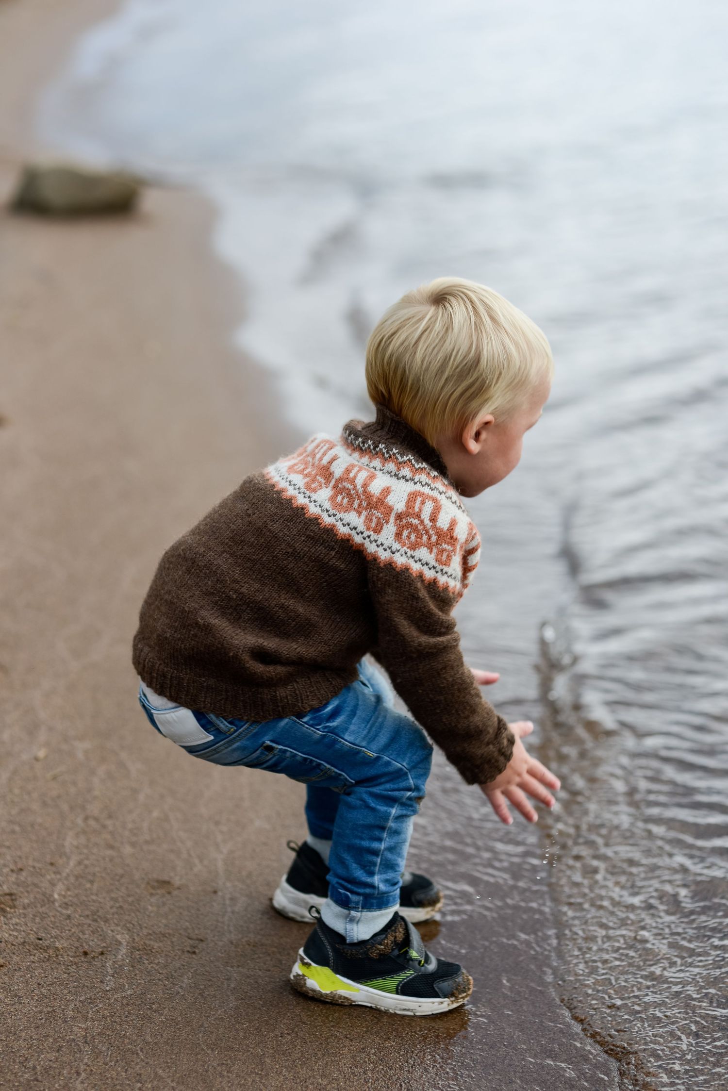 Toddler boy in brown Nordic sweater and jeans crouching at water's edge on sandy beach.