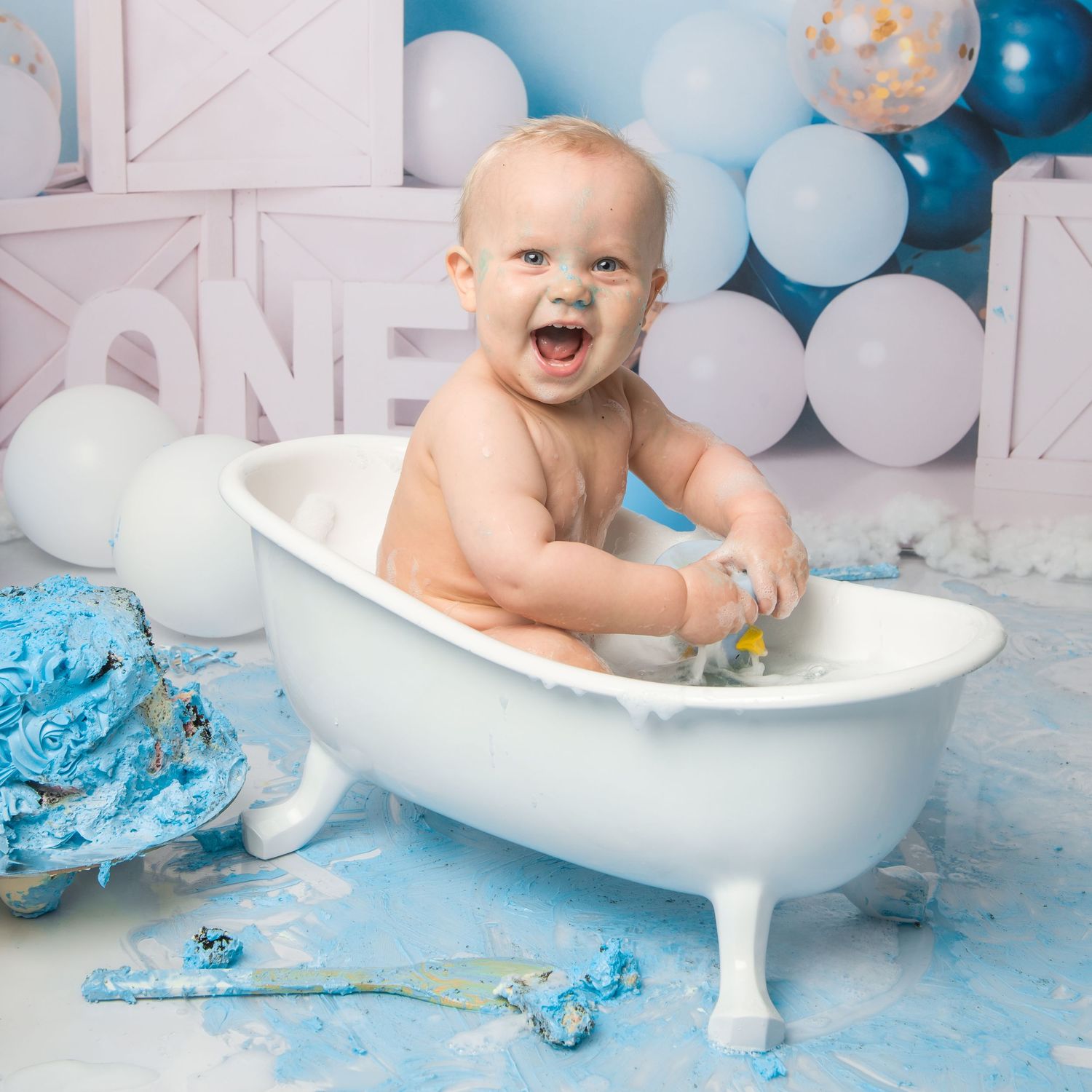 Baby sitting in white bathtub during blue themed cake smash cleanup photo session.