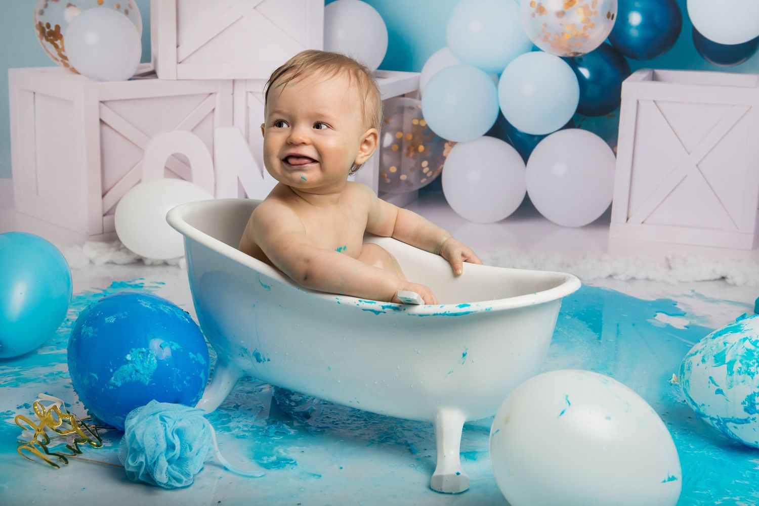 A playful cake smash photography scene with a white bathtub and blue balloon decorations.