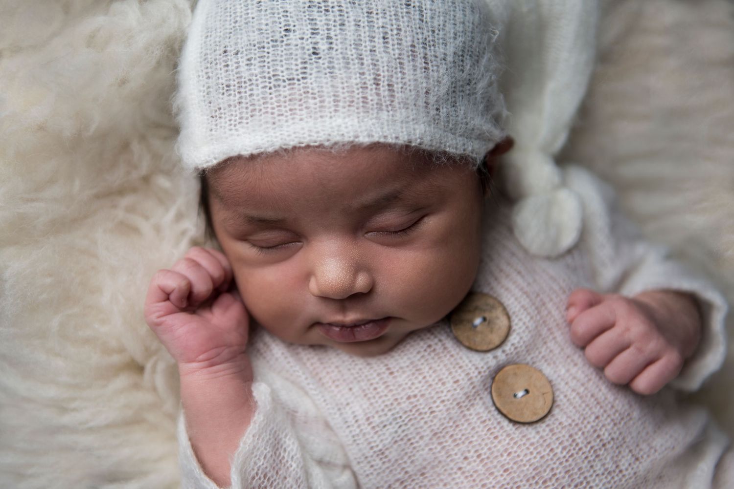 Peaceful sleeping newborn in white knit outfit with decorative buttons on furry backdrop.