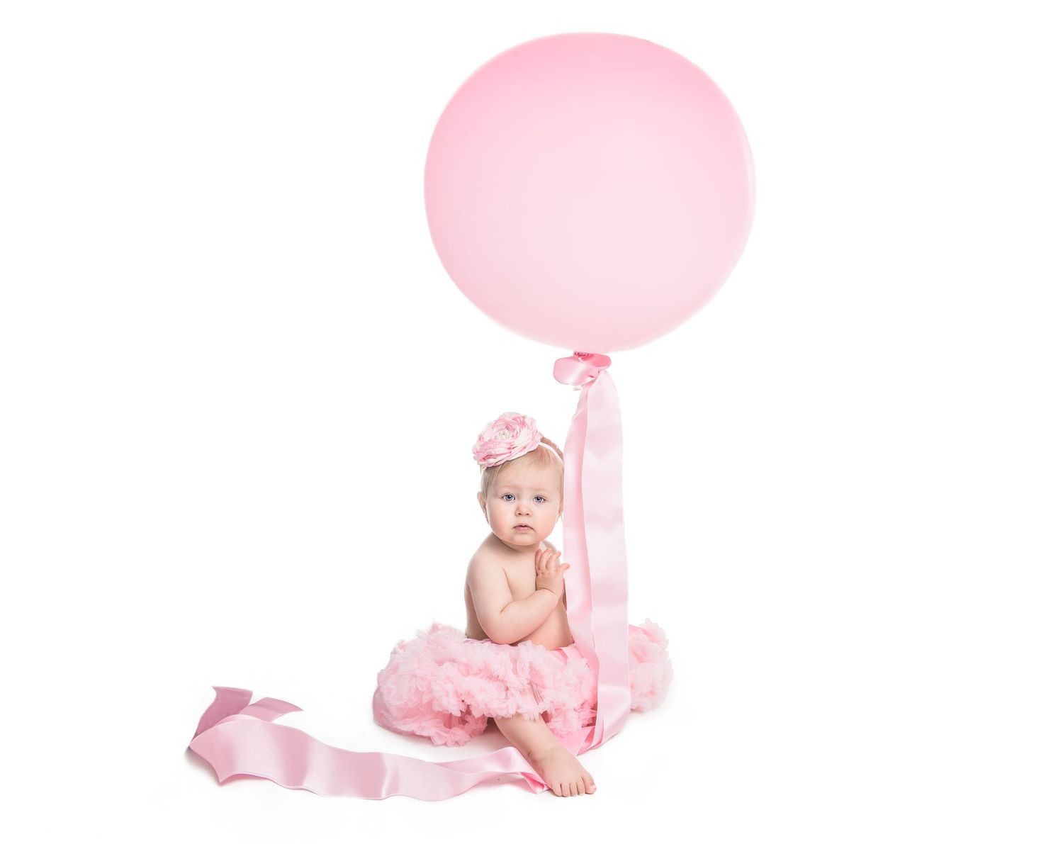 En bebis i en rosa tyllklänning sitter på ett trägolv med rosa ballonger och blomdekorationer för en födelsedagsfotografering.