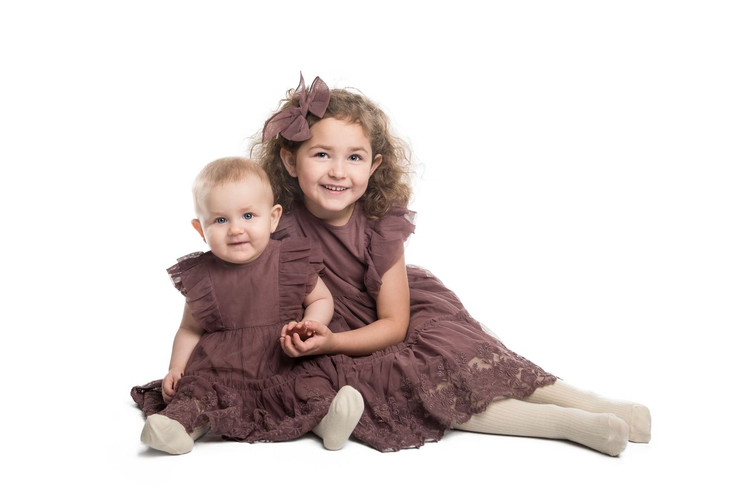 Two young children wearing matching brown ruffled dresses sit together on white background.