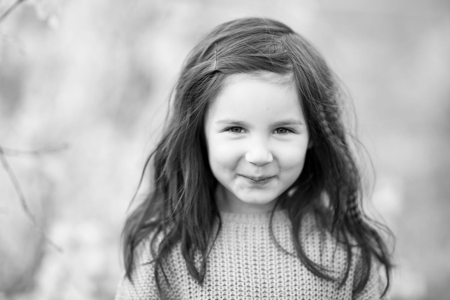 Black and white portrait of a young girl with long hair wearing a sweater, smiling softly outdoors.