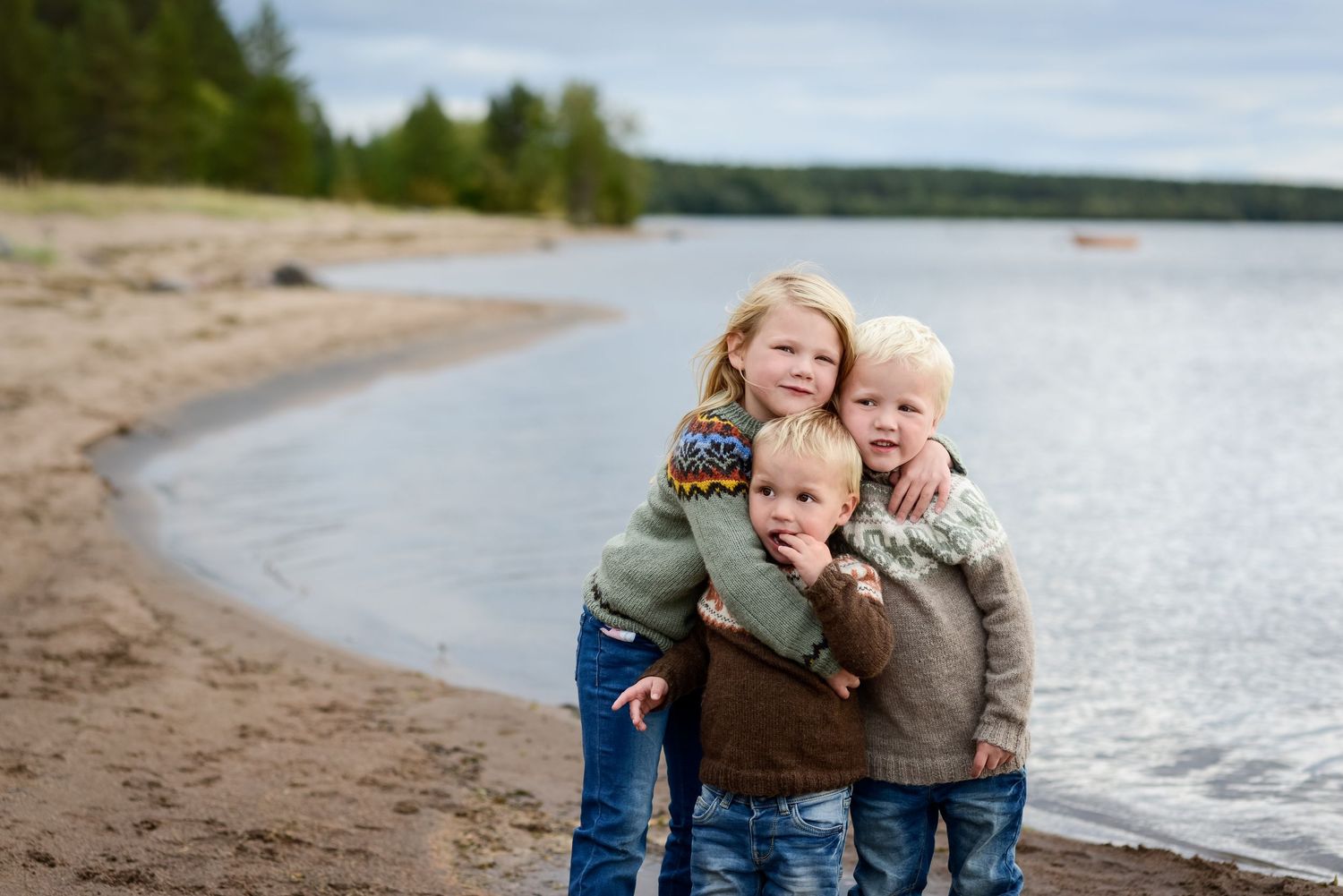 Three young blonde children hugging and smiling together on a sandy beach shoreline on a cloudy day.