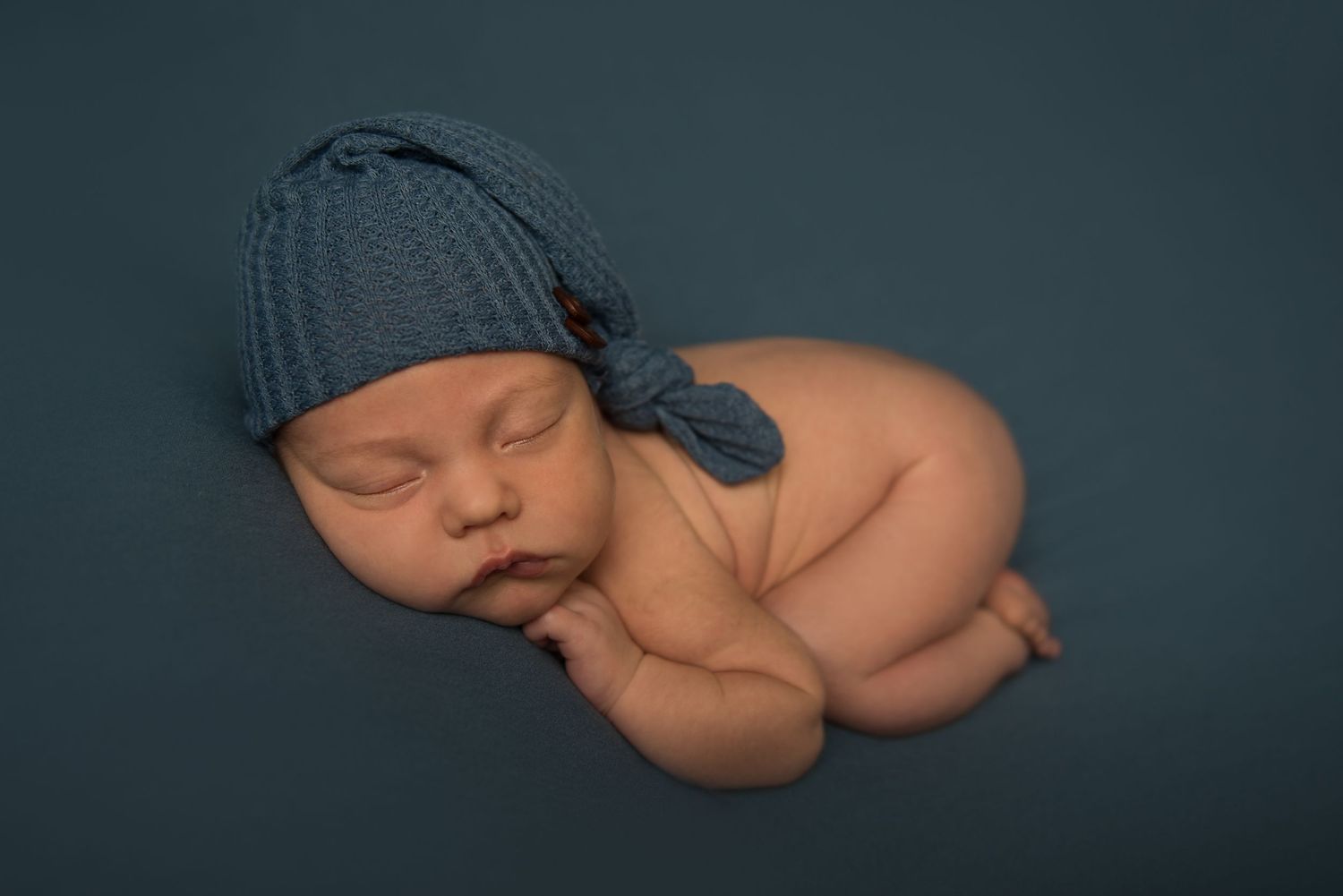 Sleeping newborn in blue knit cap posed against deep teal background.