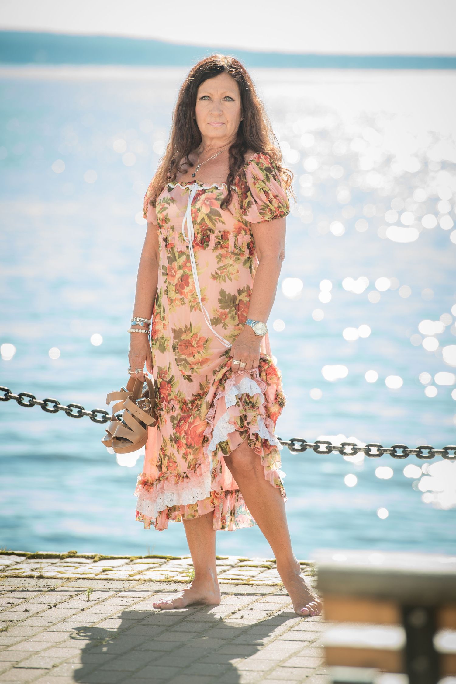 Woman in floral dress standing on seaside promenade with sparkling blue water in background on sunny day.