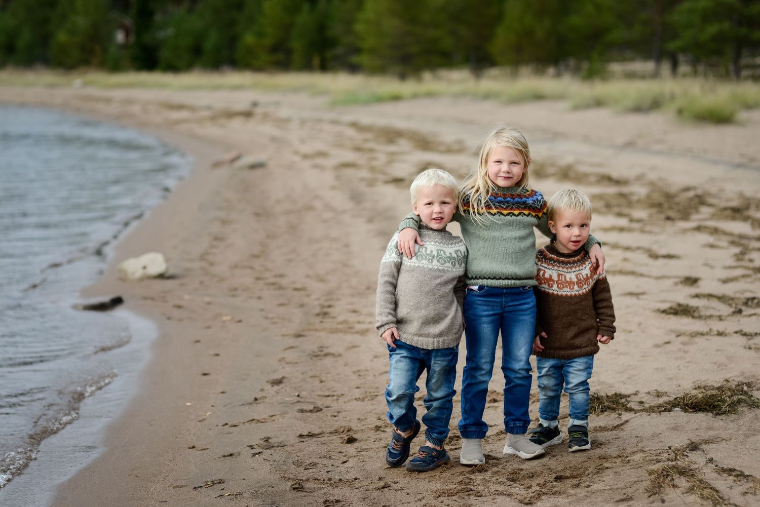 Three children in sweaters and jeans hugging on a sandy beach shore with forest in the background.