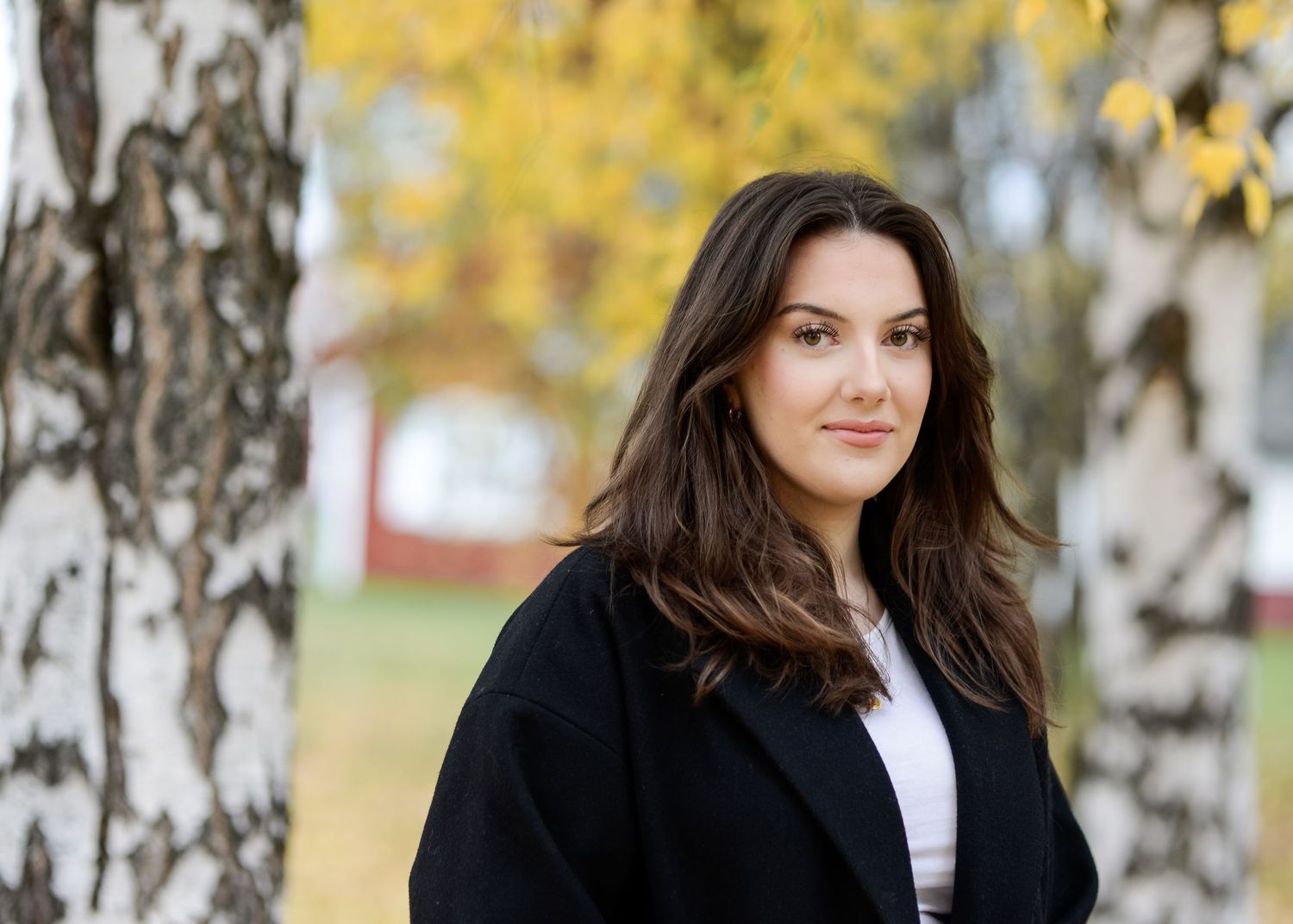 Young woman with dark hair in black coat standing among birch trees in autumn park.