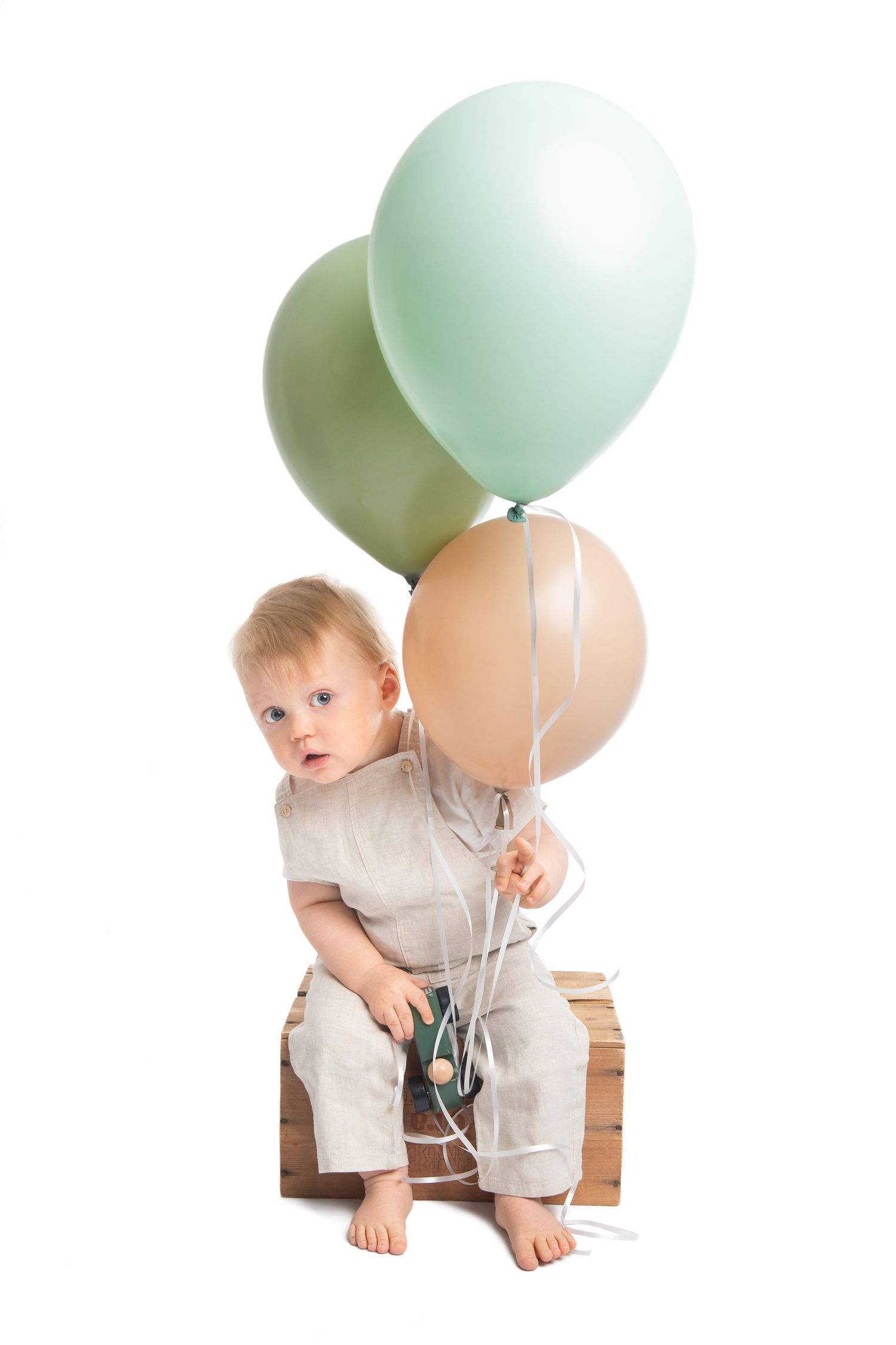 A cute toddler sits on a wooden crate holding three pastel green and beige balloons against a white background.