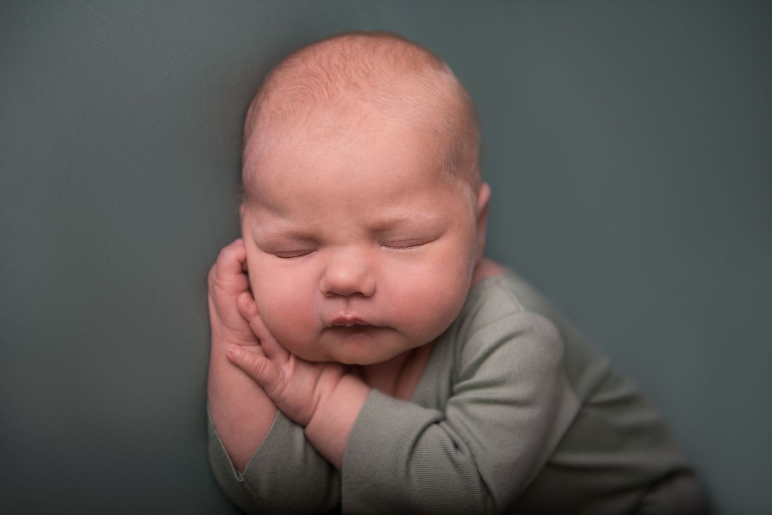 Baby wearing gray sleeper rests in curled position on teal background.