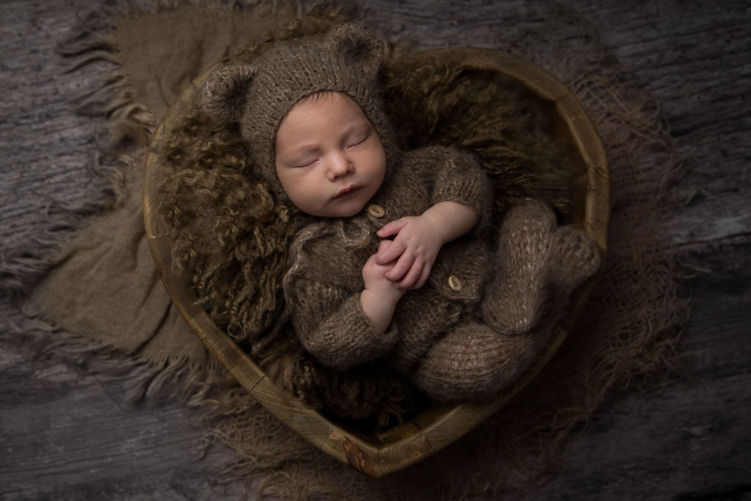 Sleeping newborn nestled in heart-shaped wooden bowl with brown knit wrap on textured gray background.